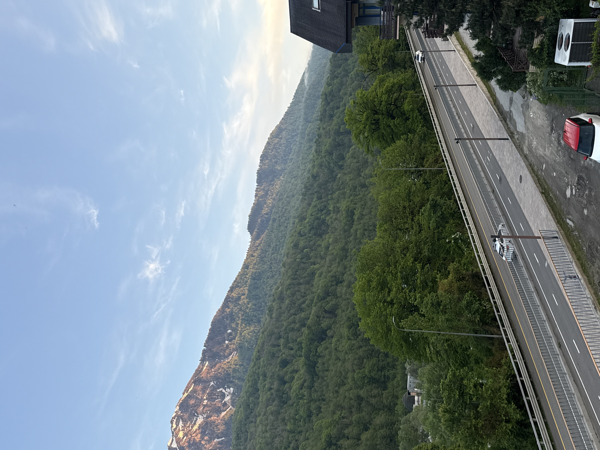 A daytime, slightly elevated view of a highway running adjacent to a steep, heavily forested mountainside. The location is Krasnaya Polyana, Russia. The sky is pale blue with scattered wispy clouds, and some haze is apparent near the top of the mountain.
The highway has multiple lanes and is bordered by a concrete median and a guardrail. There are lamp posts situated along the highway's edge. Several vehicles are visible on the road, including a white car and a white truck.
The mountainside is covered in dense green foliage, with some exposed rock and snow patches visible near the bottom. The terrain ascends steeply from the highway. Adjacent to the road are buildings, including one with a dark-colored roof and a red and white car parked nearby. A small area shows pavement with apparent discoloration.