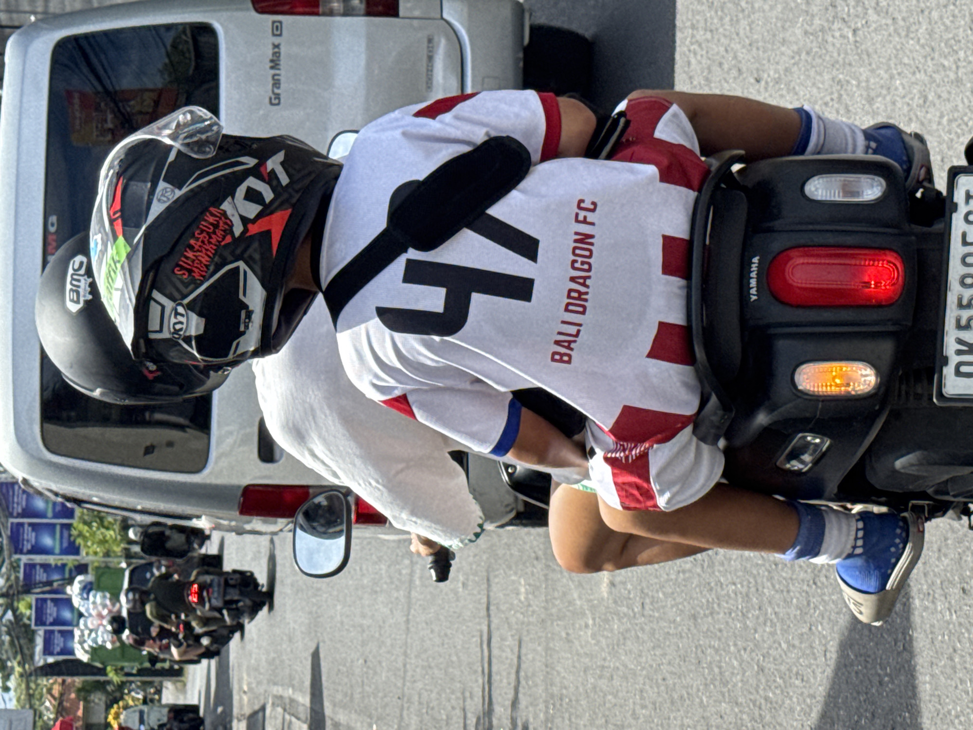 A person wearing a white Bali Dragon FC jersey with the number "47" on the back rides on a motorcycle in North Kuta, Indonesia. The individual wears a black helmet with visible brand markings and blue socks with blue and white shoes. Another person wearing a black helmet is behind the motorcycle driver. A silver van is positioned next to the motorcycle. A line of other motorcycles is visible behind the van. The street is gray asphalt. Street signs are partially visible in the background.