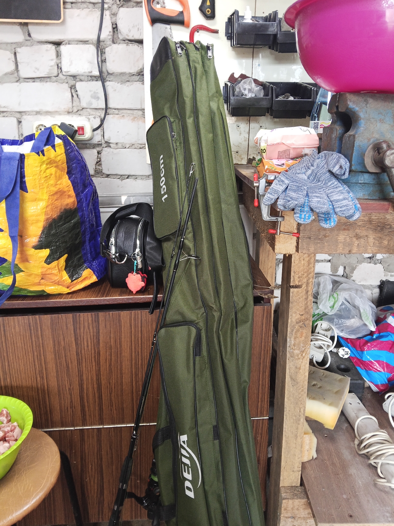 In an indoor workshop or storage area, a tall, olive-green fishing rod bag stands upright against a wooden cabinet. The bag has "DEHA 150cm" printed vertically in white text. Two fishing rods are visible extending from the top of the bag. To the left of the bag is a large, blue and yellow patterned tote bag and a small black crossbody bag with a red charm. On a wooden shelf above the cabinet, a blue glove rests on a clamp attached to a wooden plank. Various other items are present, including a pink bowl, plastic containers, wires, and bags. The wall behind the shelf is made of white brick.