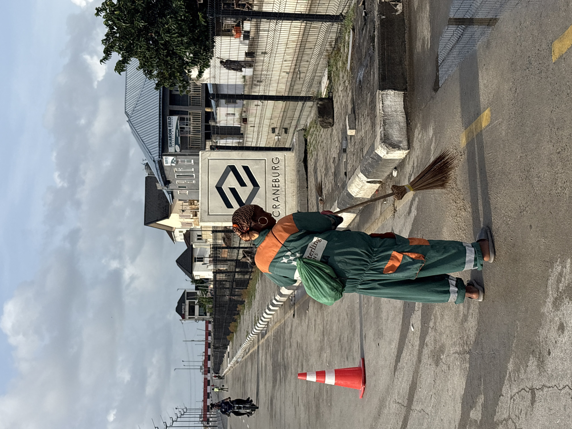 A street cleaner in a green uniform sweeps a street in Igboefon, Nigeria. The cleaner stands on a gray asphalt road near a white-and-red traffic cone and holds a broom. Behind the cleaner, a sign reads "CRANEBURG," and in the background, buildings and a metal fence are visible. Other street workers and motorcycles pass by on the road. The sky is overcast.