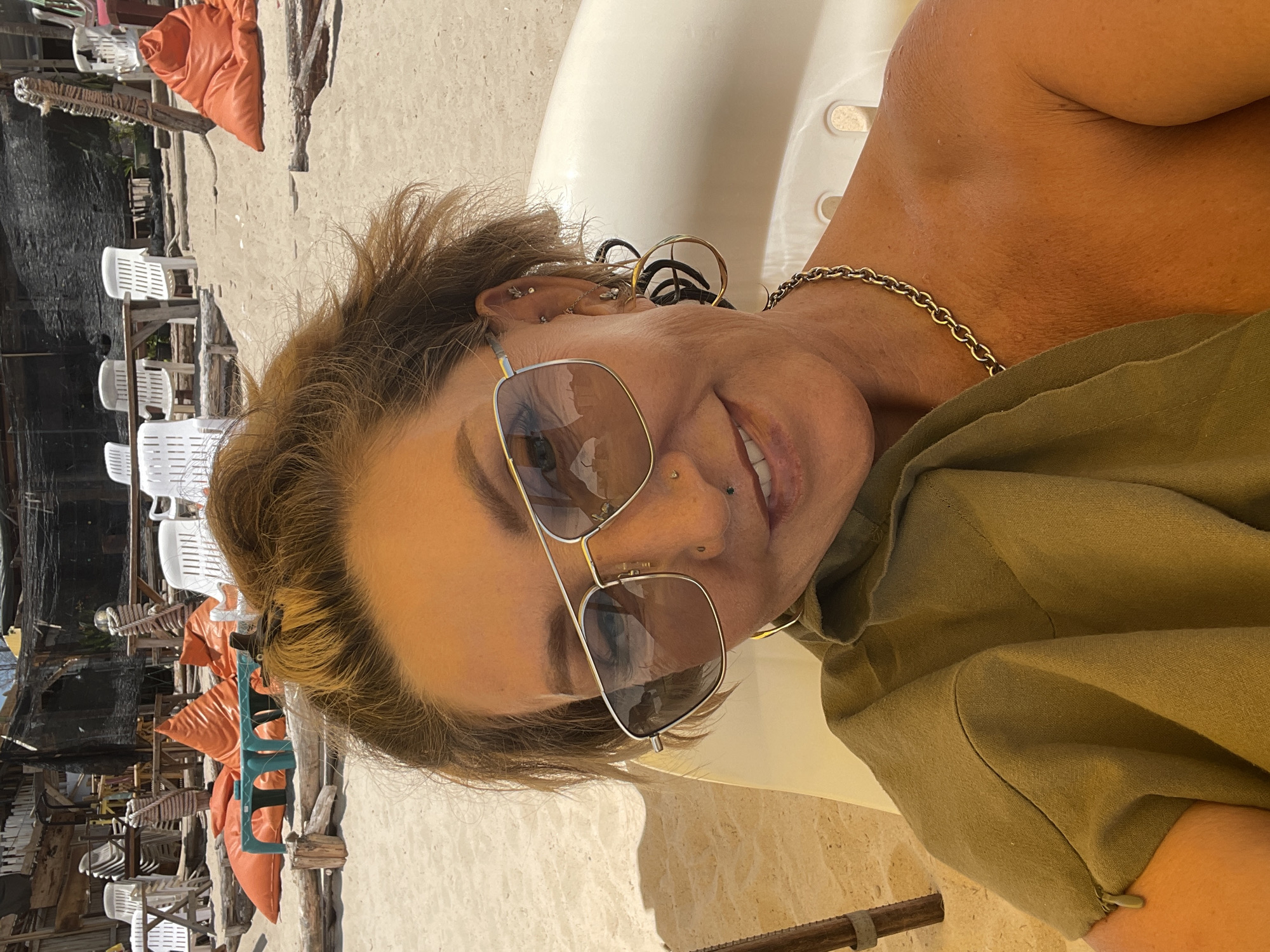 An upward-facing, rotated image captures a smiling woman relaxing in what appears to be a beachside setting in Choeng Thale, Thailand. The woman, positioned prominently in the foreground, has short, light brown hair and is wearing large aviator-style sunglasses that reflect parts of the sunny environment. Her facial expression is cheerful, with a visible smile. She sports several ear piercings on her left ear, including helix, conch, tragus, and lobe piercings, along with a philtrum (medusa) piercing and a nostril piercing on her face. A gold chain necklace is visible around her neck, and she is dressed in a loose-fitting, greenish-brown or khaki-colored top or dress. Her skin appears tanned, suggesting exposure to sun.
The background, largely behind her, depicts a casual outdoor area on sand. Scattered white plastic chairs and tables suggest a relaxed dining or lounging spot, possibly a beach bar or restaurant. Several orange, cushion-like poufs are visible on the sand among wooden structures, contributing to the rustic and informal atmosphere. Dark netting or fabric is seen further in the background, adding to the unrefined, outdoor establishment aesthetic. The bright lighting indicates it is daytime and sunny. No visible text is present in the image.
