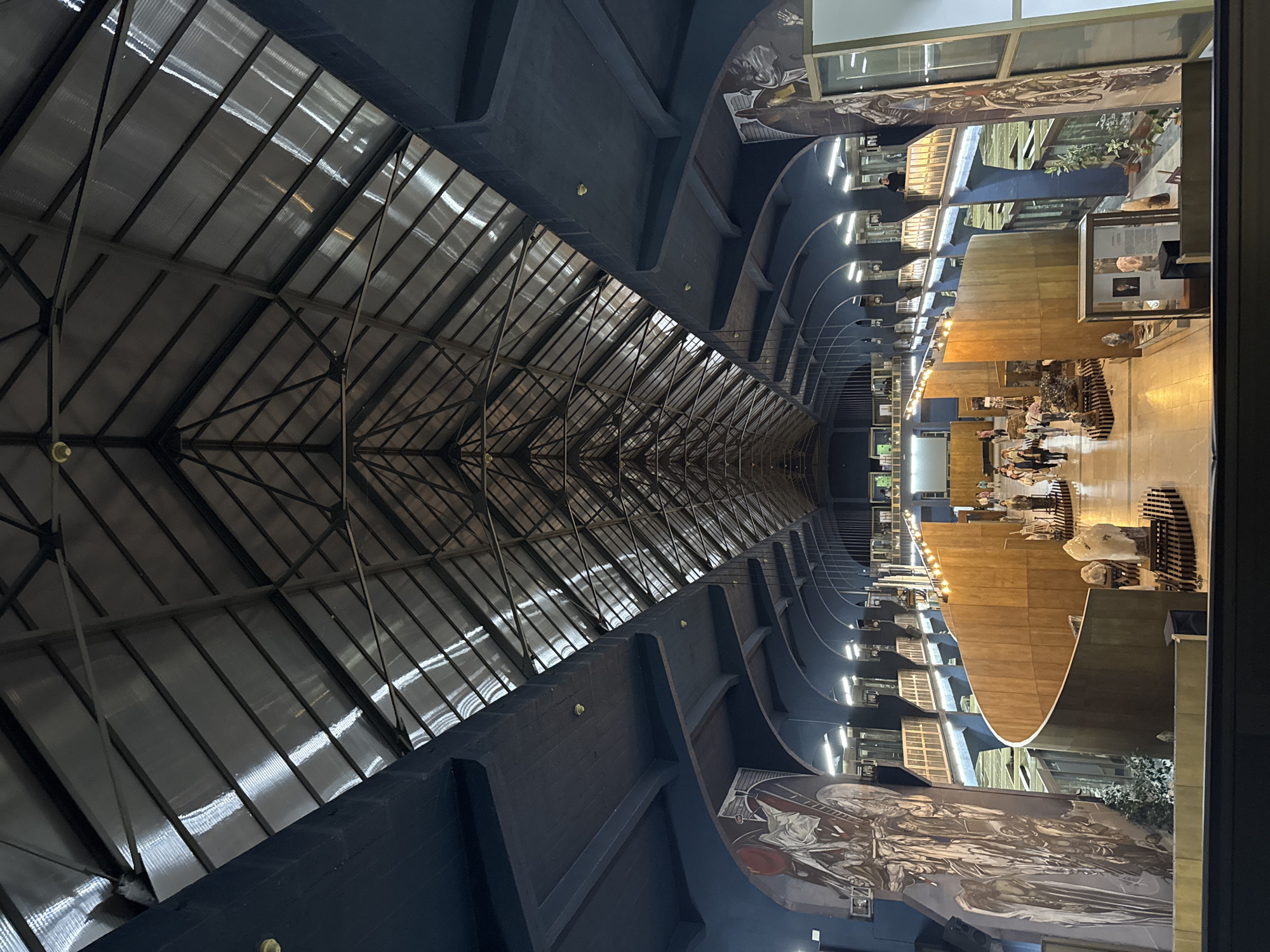 The image depicts the interior of a museum in Sofia, Bulgaria, with a distinctive architectural design. The long hall is characterized by a high, angled ceiling constructed with a network of steel beams and what appears to be translucent roofing panels, allowing natural light to filter in. The walls are dark blue and feature curved recesses, along with large murals depicting historical scenes or figures. On the right side of the frame, museum exhibits are visible, including glass display cases and wooden structures. Several people are present, walking among the exhibits. The museum has artificial lights that are illuminated throughout the building. The walls have paintings of different people.