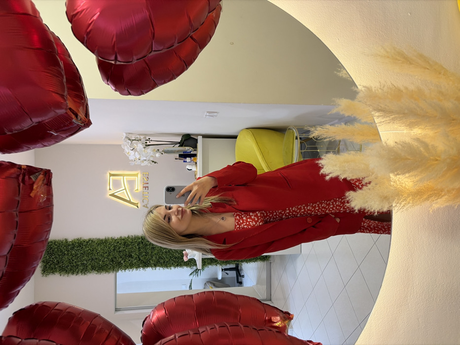 A young blonde female, approximately 20-30 years old, is depicted taking a mirror selfie inside an esthetics salon located in Budva, Montenegro. She is wearing a red long-sleeved coat over a red and white floral patterned dress, and a circular pendant necklace. Her left hand holds a grey smartphone, pointed at the mirror. The reflection shows her smiling slightly, looking at the phone screen.
The interior, primarily seen through the mirror's reflection, features white tiled flooring and white walls. A vertical panel of green artificial foliage is visible on a far wall. Above a white counter displaying various cosmetic bottles and a white orchid arrangement, a golden, illuminated sign reads "EA ESTETICA" in reverse due to the reflection. To the right of the woman's reflection, a yellow upholstered armchair with a curved back is positioned next to a small, gold-framed side table with a glass top. In the upper part of the frame, several large, red, heart-shaped foil balloons are suspended. The mirror itself is large, round, and backlit, with a cluster of dried pampas grass plumes arranged along its lower edge.