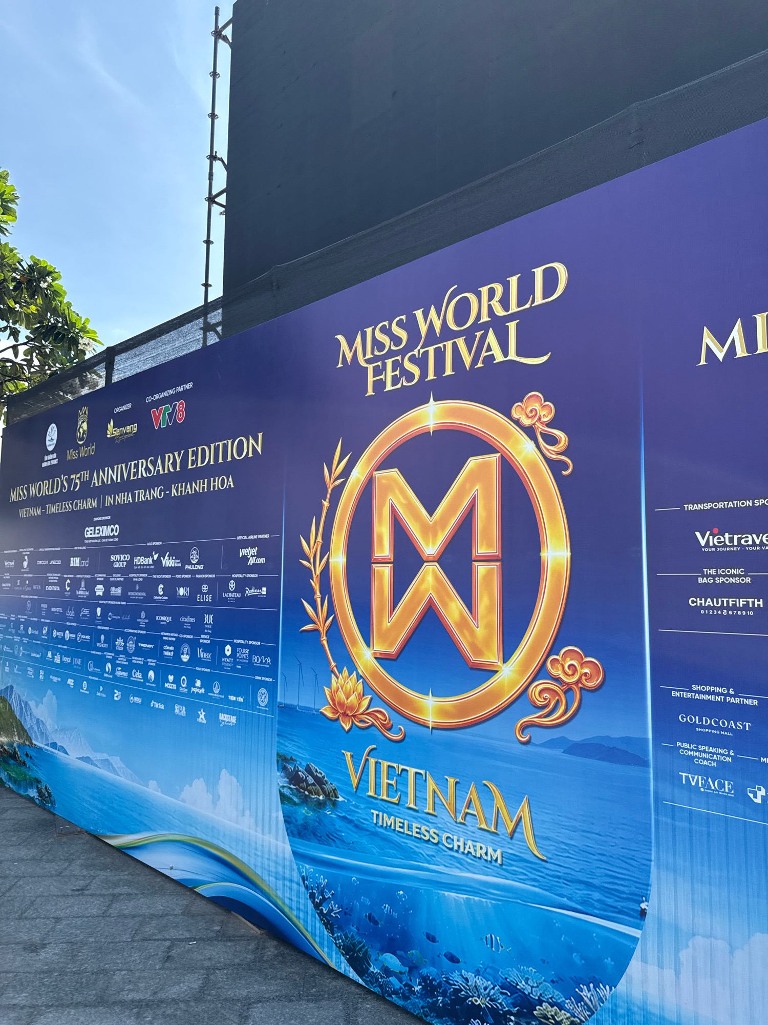 This image shows a large, blue banner advertising the "Miss World Festival" and "Miss World's 75th Anniversary Edition" in Vietnam. The banner features a large, golden "M" logo with decorative elements and the words "VIETNAM - TIMELESS CHARM". Various sponsor logos are displayed along the bottom and side of the banner. The banner appears to be set up outdoors, possibly in a public area within Nha Trang, Vietnam, as indicated by the text. The background of the banner depicts a scenic view of the ocean, islands, and underwater marine life. The lighting suggests it is daytime. There are no people or visible actions in the image.