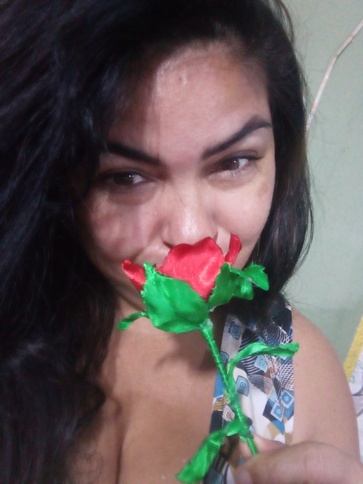 A person with dark hair and light skin is holding a red and green artificial rose up to their nose. The person is wearing a top with a geometric pattern in blue, white, and brown. The background is a pale green wall with an electrical wire running vertically. The specific location within the city of Juan Griego, Venezuela, cannot be determined from the provided image.