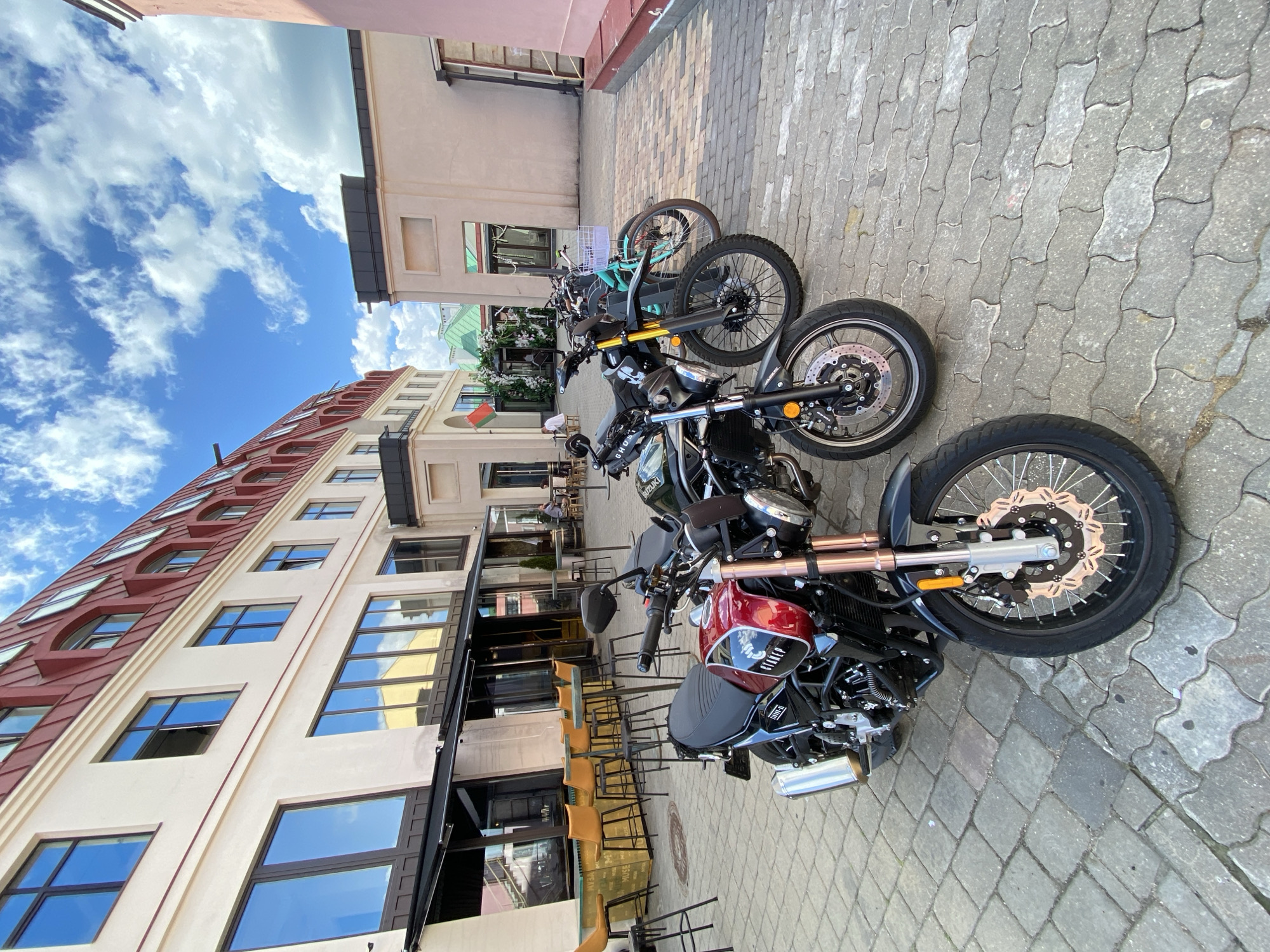 This media file shows a row of parked motorcycles and bicycles on a cobblestone street in front of buildings, likely a cafe or restaurant. The scene is outdoors during the daytime, with a bright blue sky and scattered white clouds suggesting pleasant weather.
The subjects are primarily the vehicles: at least three motorcycles and one bicycle are visible, parked in a line. One motorcycle in the foreground is dark colored with a red gas tank and copper-colored accents on the front forks. Another motorcycle behind it is dark green. The bicycle, a lighter teal color, is parked further back. There are chairs and tables set up outside the buildings, suggesting this is a public area. A few people can be seen in the background, seated at tables.
The buildings appear to be modern with large windows. One building has a distinctive red roof with curved sections. The cobblestone street provides a visual texture to the ground.
Notable details include the branding on the motorcycles. The foreground motorcycle has "SUZUKI" visible on its engine and "GTNEP" on the red tank. The overall mood is calm and relaxed, consistent with a leisurely outdoor setting. The location context provided specifies Minsk, Belarus.