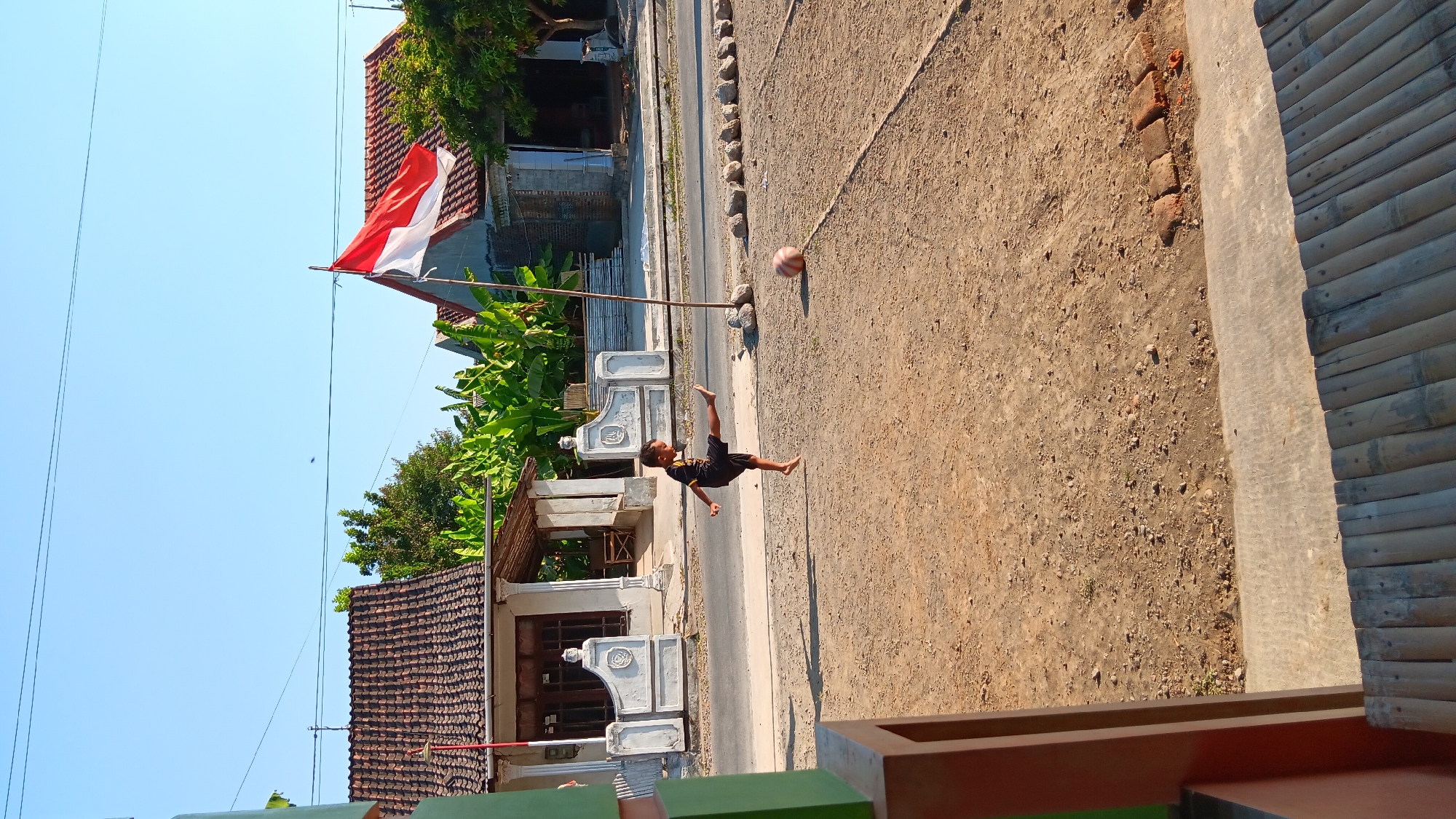In a rural Indonesian village, likely in Sonobekel, a young boy in a black t-shirt and shorts is captured mid-action, kicking a red and white striped ball. He is barefoot and positioned in a sandy, outdoor area with a dusty ground. Behind him, a dirt road runs parallel to a row of simple houses with tiled roofs. To the left of the road, a house with a white facade and decorative pillars is visible, while to the right, banana trees and other foliage frame the scene. A tall flagpole stands near the ball, flying the Indonesian flag, its red and white colors stark against the clear blue sky. The overall atmosphere is one of everyday life in a village setting.