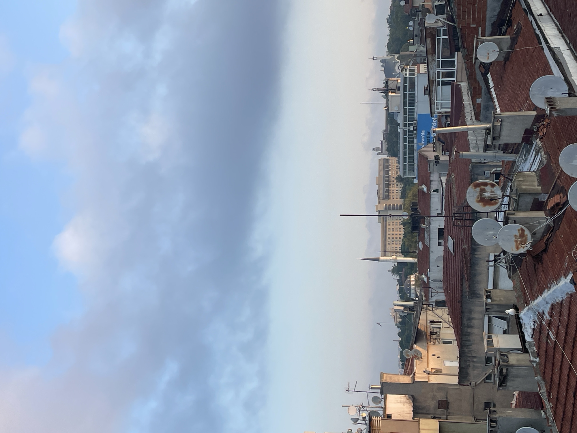 The image depicts a rooftop view of Kadıköy, Turkey, under a cloudy sky. In the foreground, several buildings with red-tiled roofs and satellite dishes are visible. A solitary seagull stands on one of the rooftops. In the middle ground, more buildings and structures are present, including what appears to be a mosque with a tall minaret. Further in the background, larger buildings and distant hills can be seen. The overall scene suggests an urban environment during daylight hours, possibly early morning or late afternoon, given the soft lighting.