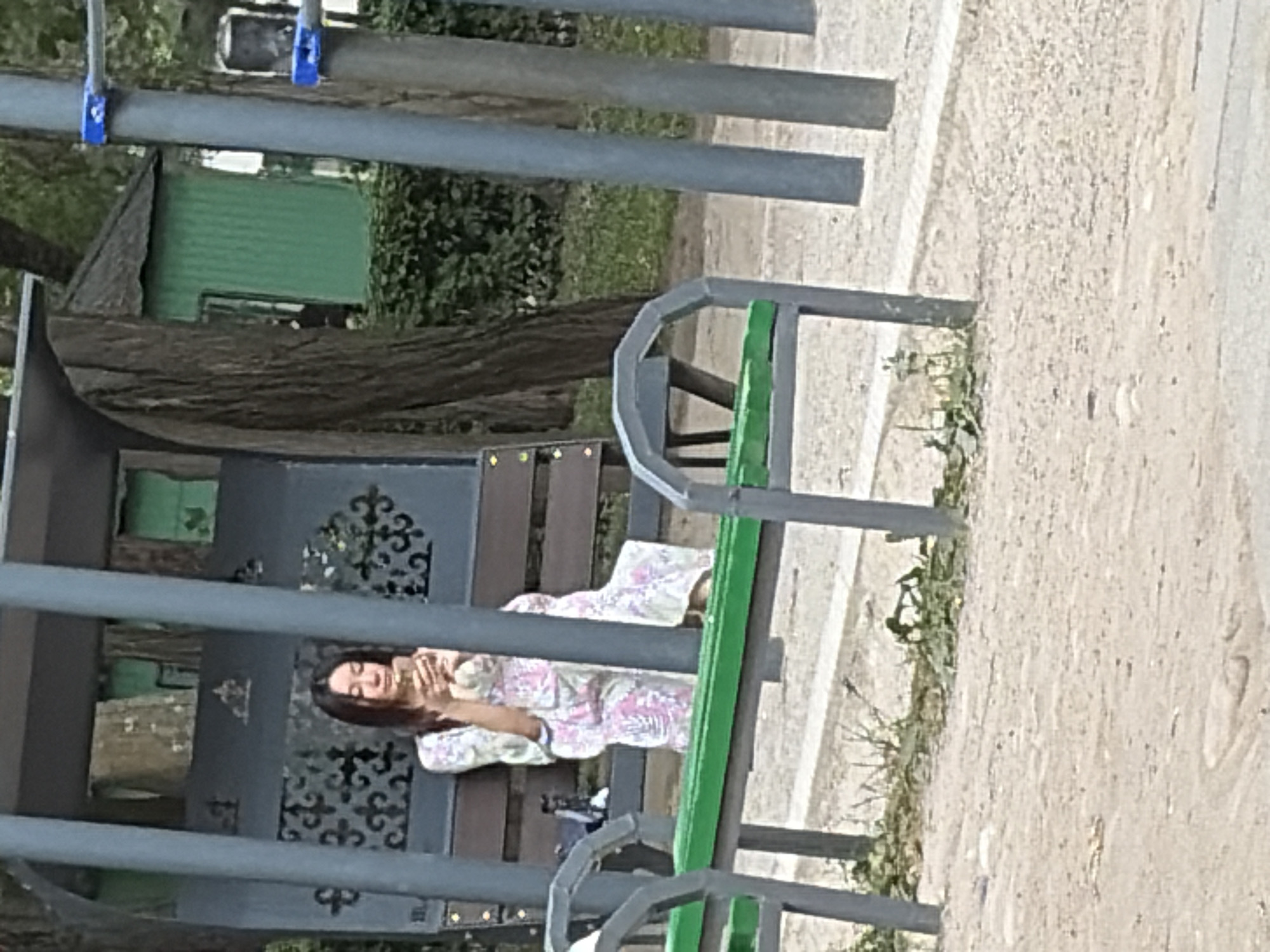 A woman sits on a bench under a covered shelter and looks at her phone. She is wearing a light-colored dress with a floral pattern. A green building is visible in the background. The scene is outdoors in what appears to be a park or playground.