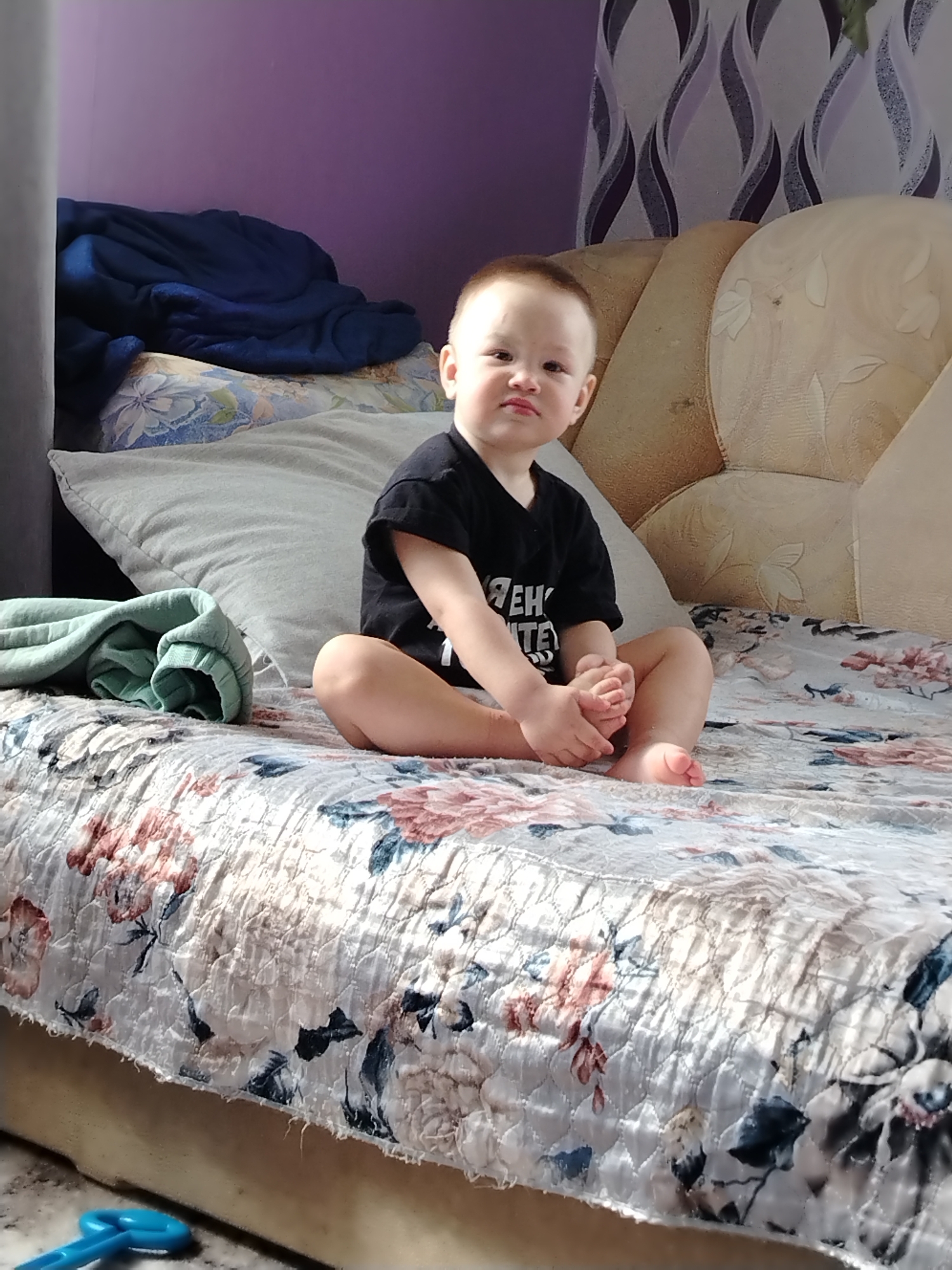 A young child sits cross-legged on a bed. The child is wearing a black t-shirt and is holding their feet. The bed has a floral patterned quilt. Behind the child is a pillow and a headboard. A blue blanket is on the left. A pair of green sweatpants are also on the bed. In the bottom left corner, a blue plastic toy is visible on the floor. The room has purple walls and patterned curtains. The location is Гожан, Russia.