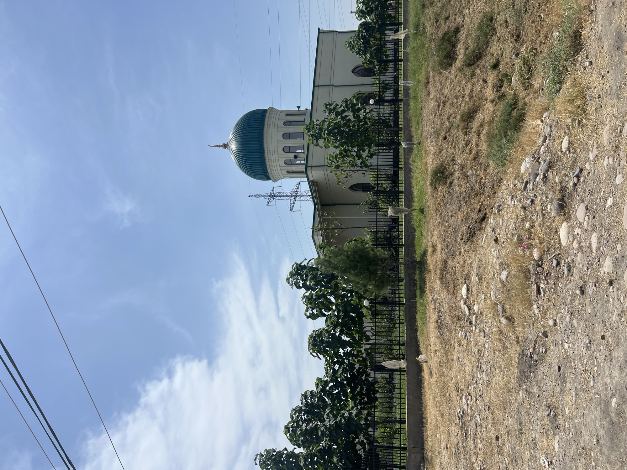 The image shows a mosque with a green dome, located in Karamanas, Uzbekistan. The mosque is a light-colored building with arched windows and a prominent dome topped with a crescent. It is surrounded by a black metal fence and lush green trees. In the foreground, the ground is dry and rocky, with sparse vegetation. Overhead, power lines stretch across a clear blue sky with scattered clouds. A transmission tower is visible to the left of the mosque. There are no people visible in the image.