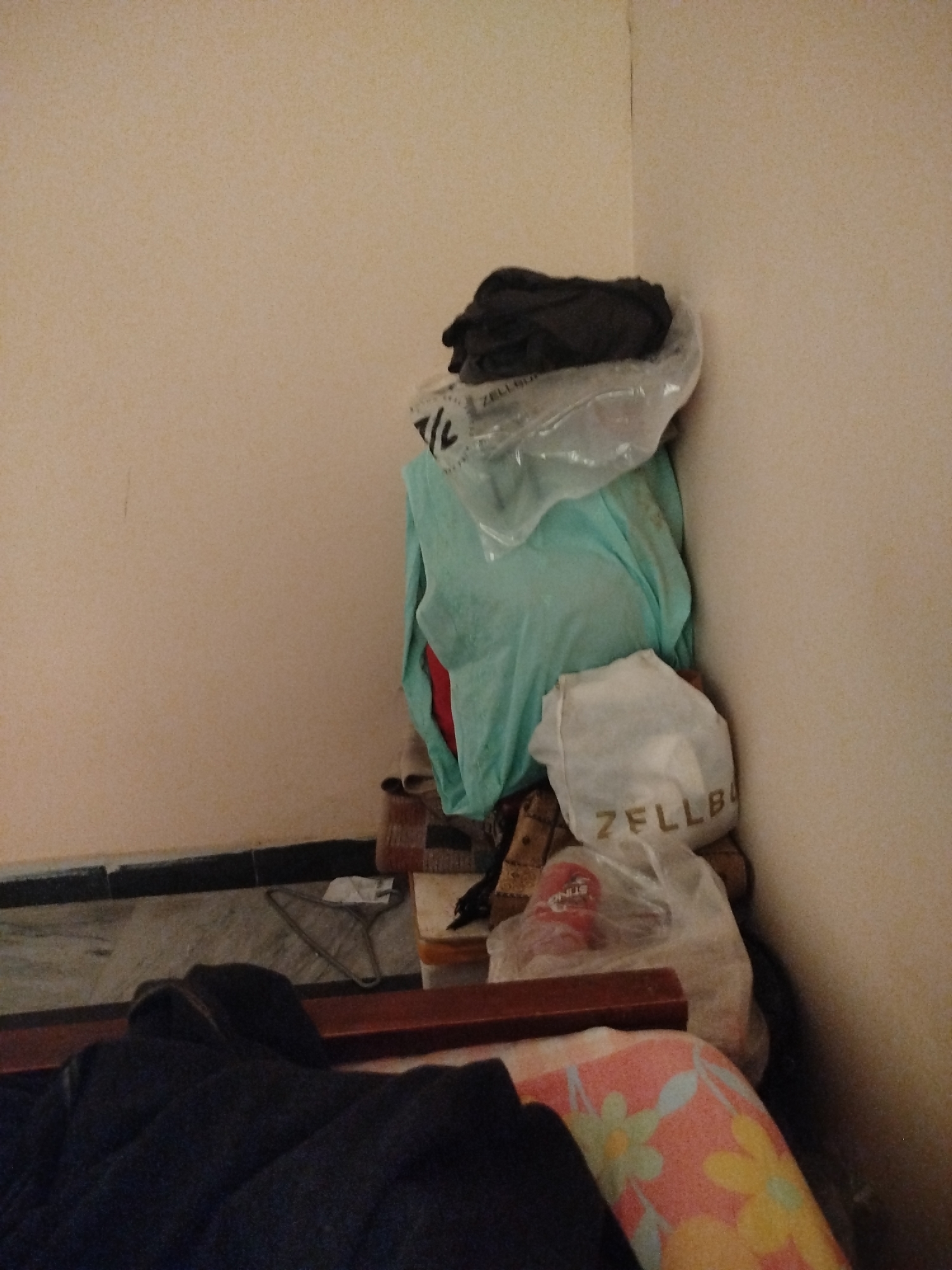 A stack of clothing is piled in the corner of a room. The clothing appears to be primarily in plastic bags. The room has cream-colored walls and a dark baseboard. In the foreground, a dark blanket is draped over what appears to be furniture. The floor is tiled with a marble pattern. There are no people visible in the image. The scene does not provide any specific information to indicate its location within the city of Lassan, Pakistan.