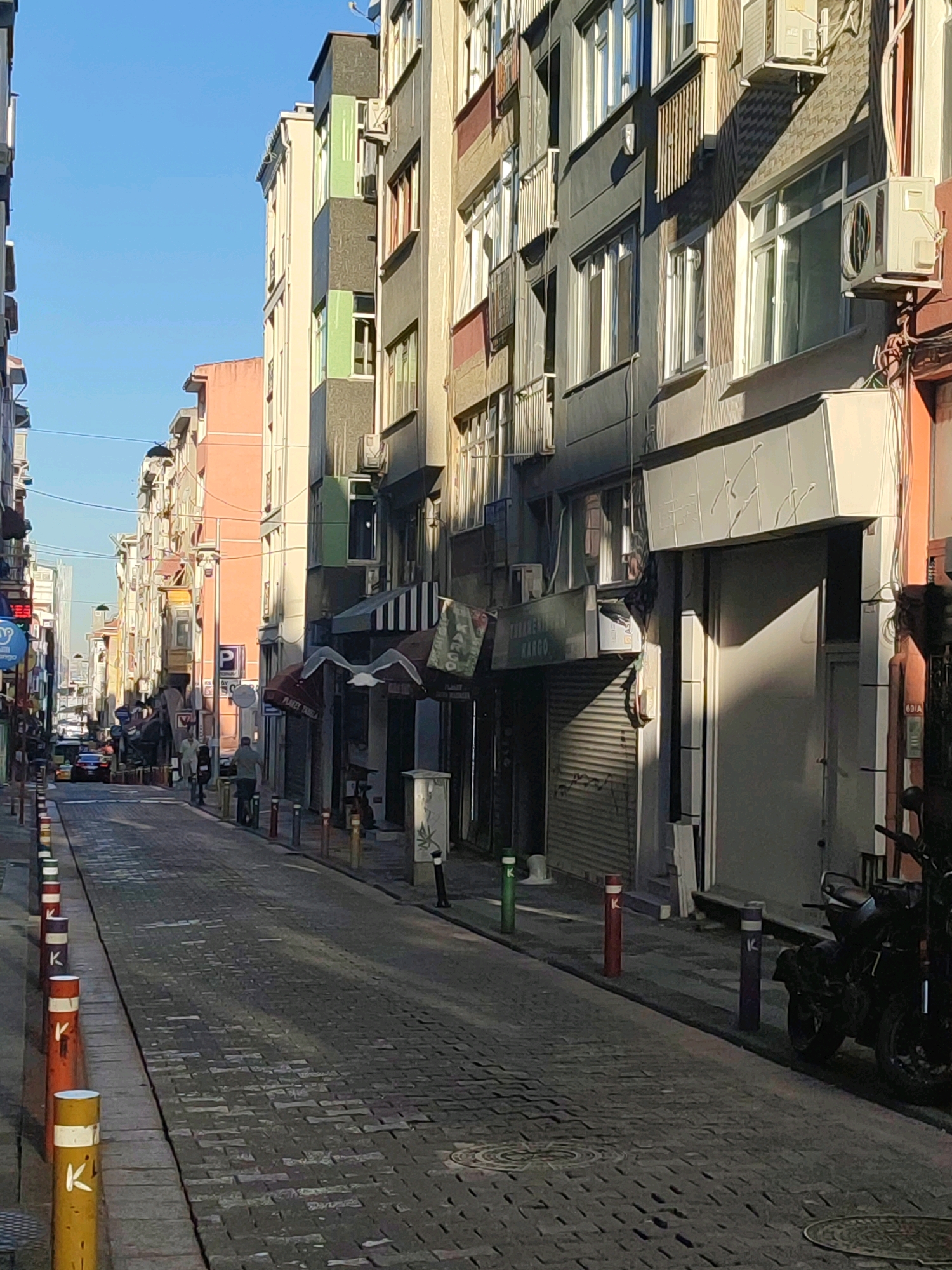 The image captures a narrow, cobblestone street in Kadıköy, Turkey, lined with multi-story buildings on both sides. The sky is clear and blue, indicating daytime with sunlight casting shadows on the street and buildings. Several parked motorcycles are visible on the right side of the street. Buildings feature a mix of architectural styles and colors, with some displaying awnings and air conditioning units. Bollards of various colors, some with white stripes and the letter "K," are placed along the sidewalks. A few individuals are present, walking along the street. Traffic appears light, with at least one car and a taxi visible in the distance. The street level of the buildings includes shops with rolled-down shutters.