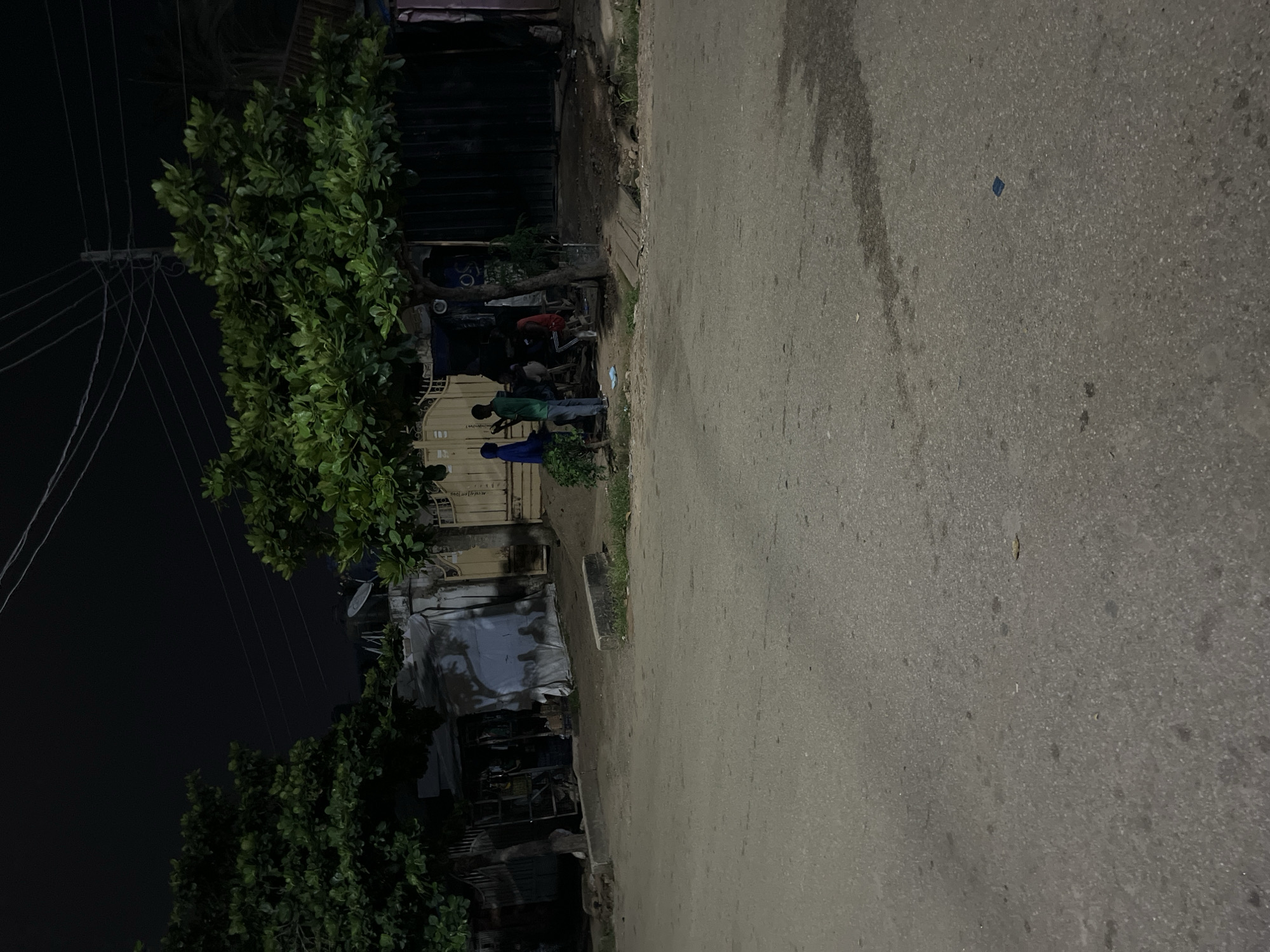 This image depicts a nighttime street scene in Abuja, Nigeria. The foreground is dominated by a paved road, with a slight sheen suggesting moisture or oil. A concrete curb runs along the side of the road, separating it from a dirt and gravel area. In the background, a building with a light-colored, possibly tarp-covered facade is visible, flanked by lush green trees. A metal gate and a darker, possibly wooden structure are also present. Several people are gathered in the area between the trees and the building. One person in a blue garment stands to the left, facing a person in a green shirt. Another individual in a red shirt is seated. The overall impression is of a common area or a roadside establishment at night. The presence of overhead wires and a utility pole suggests an urban or semi-urban environment. The lighting is dim, casting shadows and creating a somewhat grainy texture in the image.