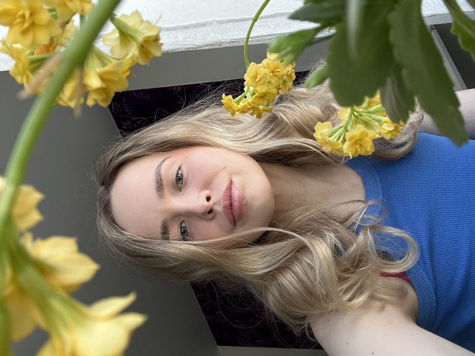 The image depicts a young adult female captured from the chest up, positioned centrally, looking directly at the camera. She has long, wavy blonde hair, light blue eyes, and light-toned skin, with a pink blush visible on her cheeks and light pink lipstick. She wears a ribbed blue tank top; a red strap, likely from an undergarment, is visible on her left shoulder. The scene is indoors, with a light-colored ceiling overhead. To her left, a dark, patterned surface, possibly wallpaper or a curtain, is visible. To her right, a vertical light-colored structure, consistent with a window frame, is present, suggesting ambient light sources from that direction. Numerous clusters of small, bright yellow flowers, along with green leaves, are prominently arranged in the foreground and mid-ground, partially framing the woman. The specific location within the city of Chaikovsky, Russia, cannot be visually confirmed from the details present in the image.