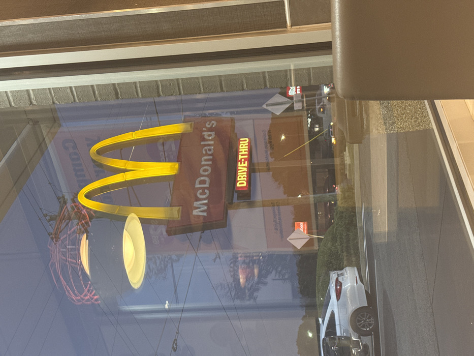 A McDonald's sign with golden arches and "DRIVE-THRU" is reflected in a window. Outside the window, a white car is parked near a green bush. Streetlights and possibly other signs are visible in the reflection. A portion of a building's interior, including a chair, is visible in the upper right corner. The scene is captured through the glass window.