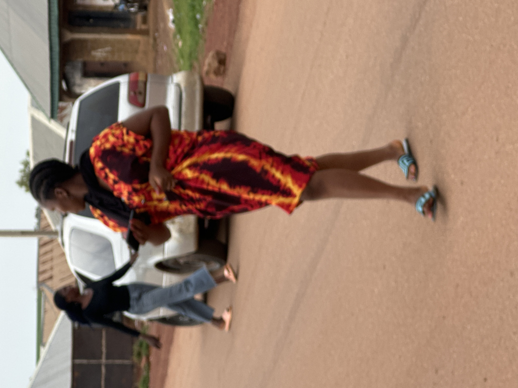In Durumi, Nigeria, a street scene unfolds. The ground is a reddish-brown dirt road. In the foreground, a woman walks away from the camera, her back and right side visible. She wears a colorful, tie-dye dress in shades of red, yellow, and black, with blue and pink sandals. She is holding a dark object, possibly a phone, in her right hand. Further back, a second woman walks across the road, her left side visible. She is wearing a black long-sleeved shirt and blue jeans, with light pink sandals. Behind the women, a silver minivan is parked or moving slowly across the road. In the background, there are buildings with corrugated metal roofs, characteristic of the area. The sky is overcast, suggesting a cloudy day. The overall impression is of a typical day in a Nigerian community.