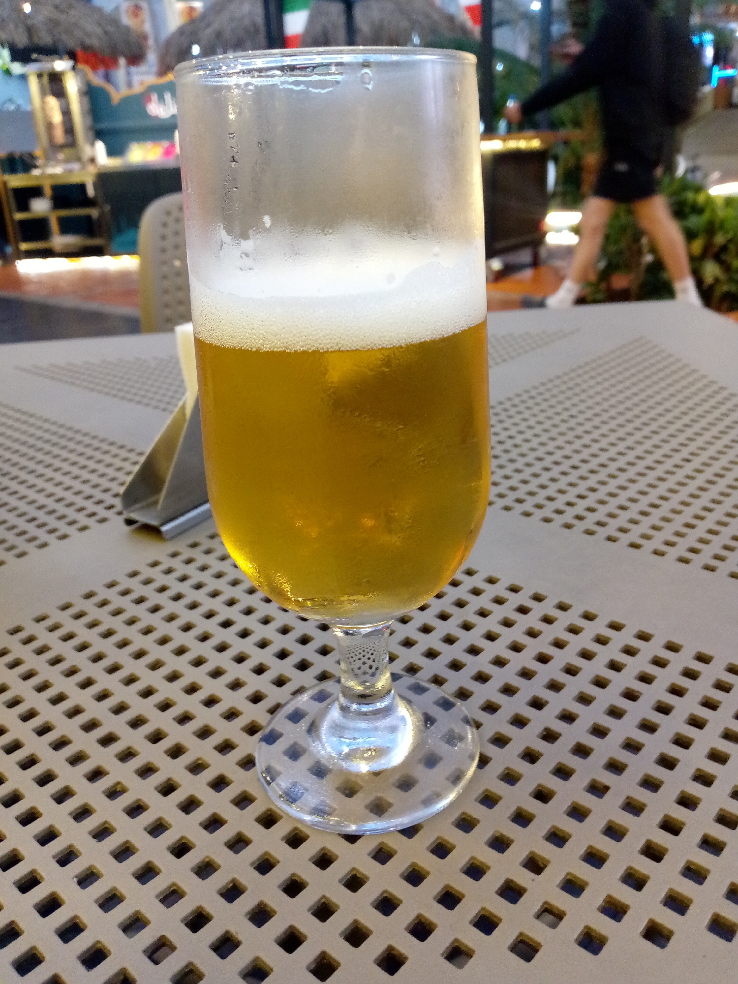 A beer glass filled with a light-colored liquid and a foamy head sits on a gray plastic table with a grid pattern. A metal holder is partially visible to the left of the glass. In the blurred background, there are indistinct shapes resembling tiki huts, possibly indicating an outdoor bar or restaurant setting. A person is walking away in the distance. The location appears to be outdoors, possibly in an urban area.