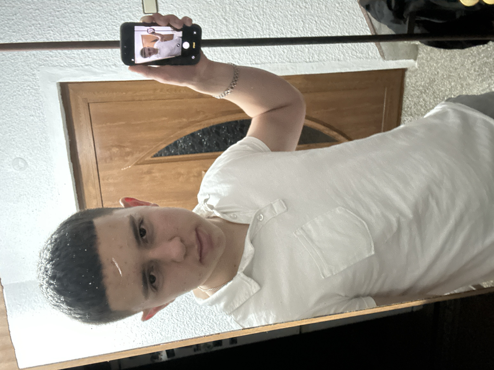 A person with short, dark hair is taking a selfie in a mirror. The person is wearing a white shirt and a silver necklace. The person is holding a black smartphone in their left hand, displaying an image of themself. The background includes a brown wooden door with a decorative glass panel. The wall behind the door has a textured, light-colored surface. A dark-colored rod or bar runs horizontally across the top of the image. Part of a carpet and some clothes are visible in the upper right corner. The image appears to be taken indoors, possibly in a residential setting. The location in the city of Zlatograd, Bulgaria, is not evident from the image.