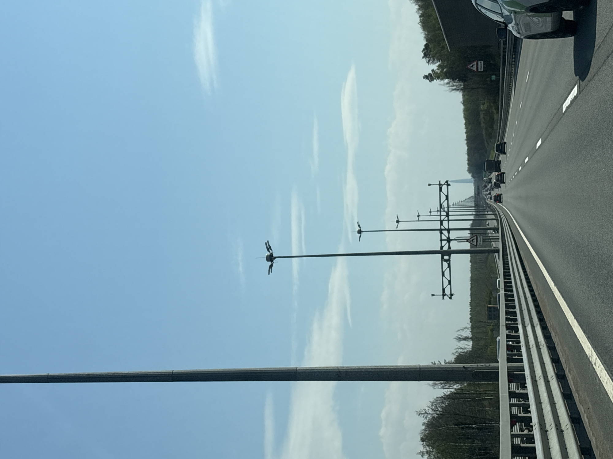 The image shows a highway in Pesochny, Russia. The view is from a vehicle on the rightmost lane, looking straight ahead. The road is lined with metal guardrails and tall light poles with anemometers. Several cars and trucks are visible in the distance. The sky is blue with scattered white clouds. The road is surrounded by trees.