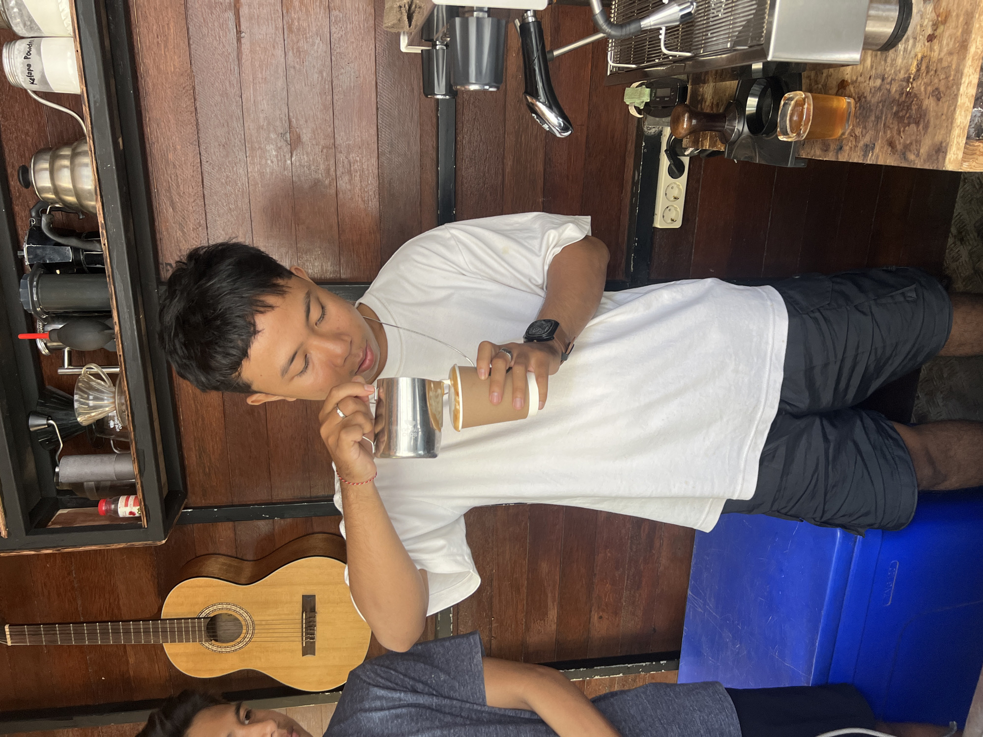 A young man is in the process of making coffee, holding a metal pitcher in his left hand and a paper cup in his right. He appears to be pouring milk from the pitcher into the cup. He is wearing a white t-shirt and black shorts. A second person, only their torso and head visible, is seated in the foreground.
The scene is indoors, with wooden walls and floorboards. Various coffee-making equipment is visible, including an espresso machine on a wooden counter. There is also a guitar resting on the floor. Shelves on the left hold jars and other containers, one of which has "Kelapa Pouch" written on it. The overall impression is of a casual, possibly home or small cafe setting. The lighting suggests it could be daytime, but there are no clear indicators of the time of day or weather.