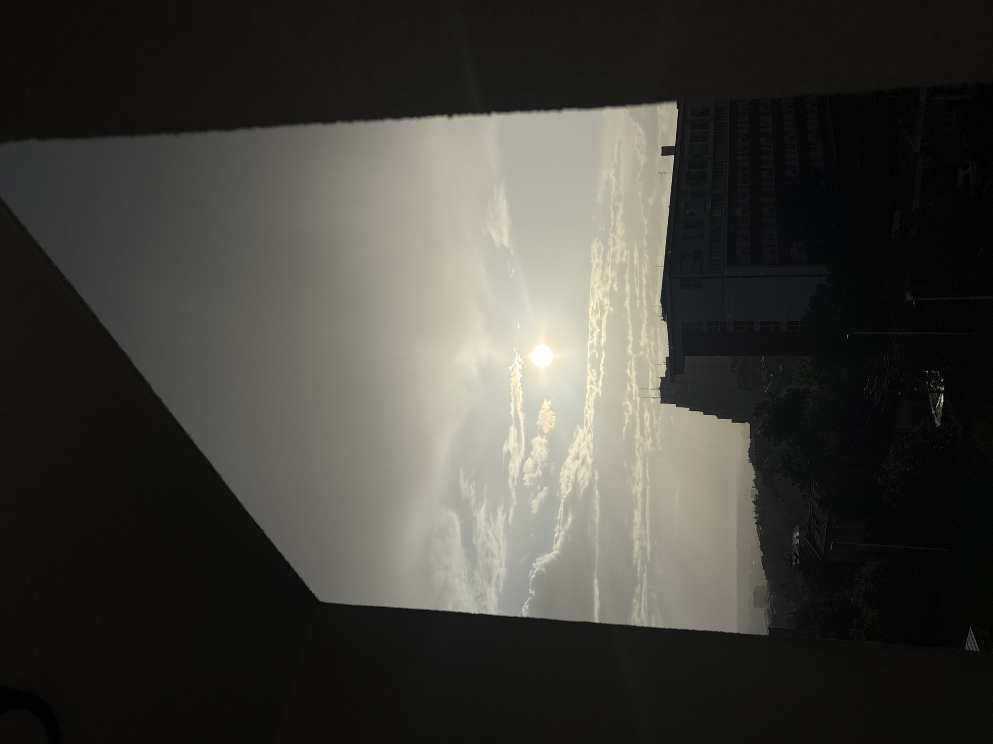 The image captures a view framed by a window, likely from inside a building in Belgrade, Serbia. The sun is visible through a layer of clouds, with a bright glare obscuring details. A tall building is located on the right side of the frame with multiple windows that form a pattern. The view extends towards a hazy horizon, and a tree line can be discerned in the distance. The window frame is dark and obscures a significant portion of the view, creating a sense of looking out from a darkened room. No people are visible in the image.