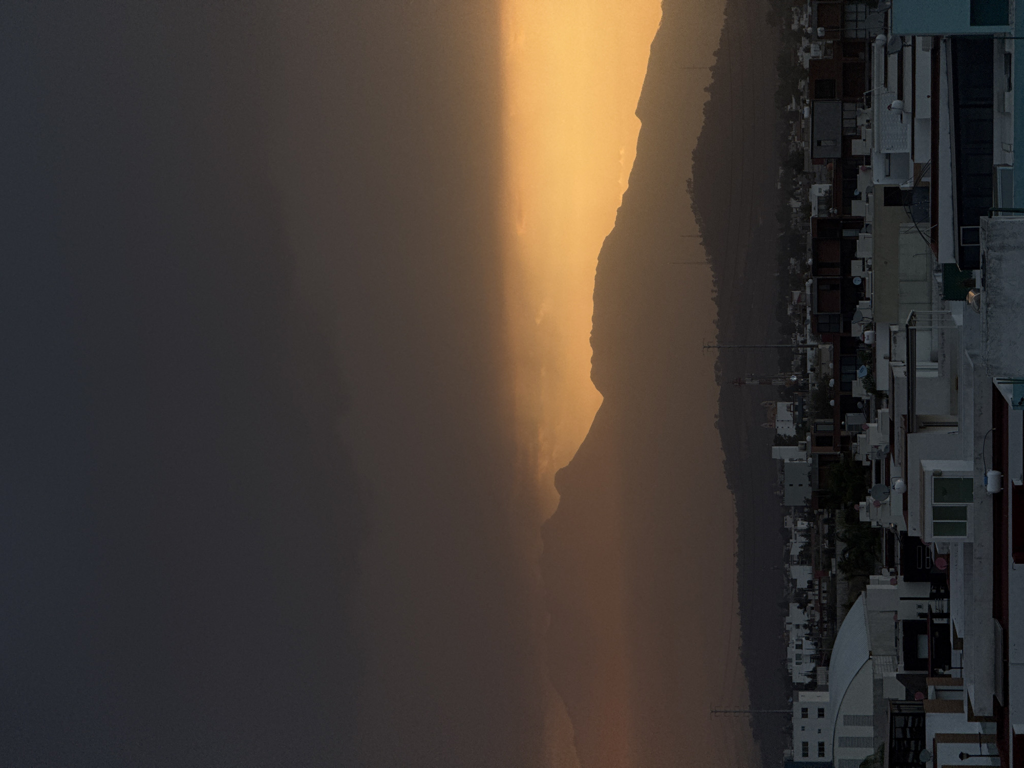 The image depicts an urban environment in San Bernardino Tlaxcalancingo, Mexico, at twilight. The sky transitions from a dark gray, heavily clouded upper portion to a brilliant orange and yellow stratum along the horizon, where sunlight penetrates. In the midground, multiple dark, silhouetted mountain ranges rise against this brightly illuminated horizon. The foreground and lower midground are filled with a dense collection of flat-roofed buildings, predominantly light-colored, indicative of residential or commercial structures. Numerous red satellite dishes and white cylindrical water tanks are visible on various rooftops. A large building featuring a curved, white metallic roof is prominent in the lower-left foreground. Tall metal transmission towers and power lines are observable within the cityscape and against the lower slopes of the distant hills. No human figures, specific actions, or interactions are discernible in the scene.
