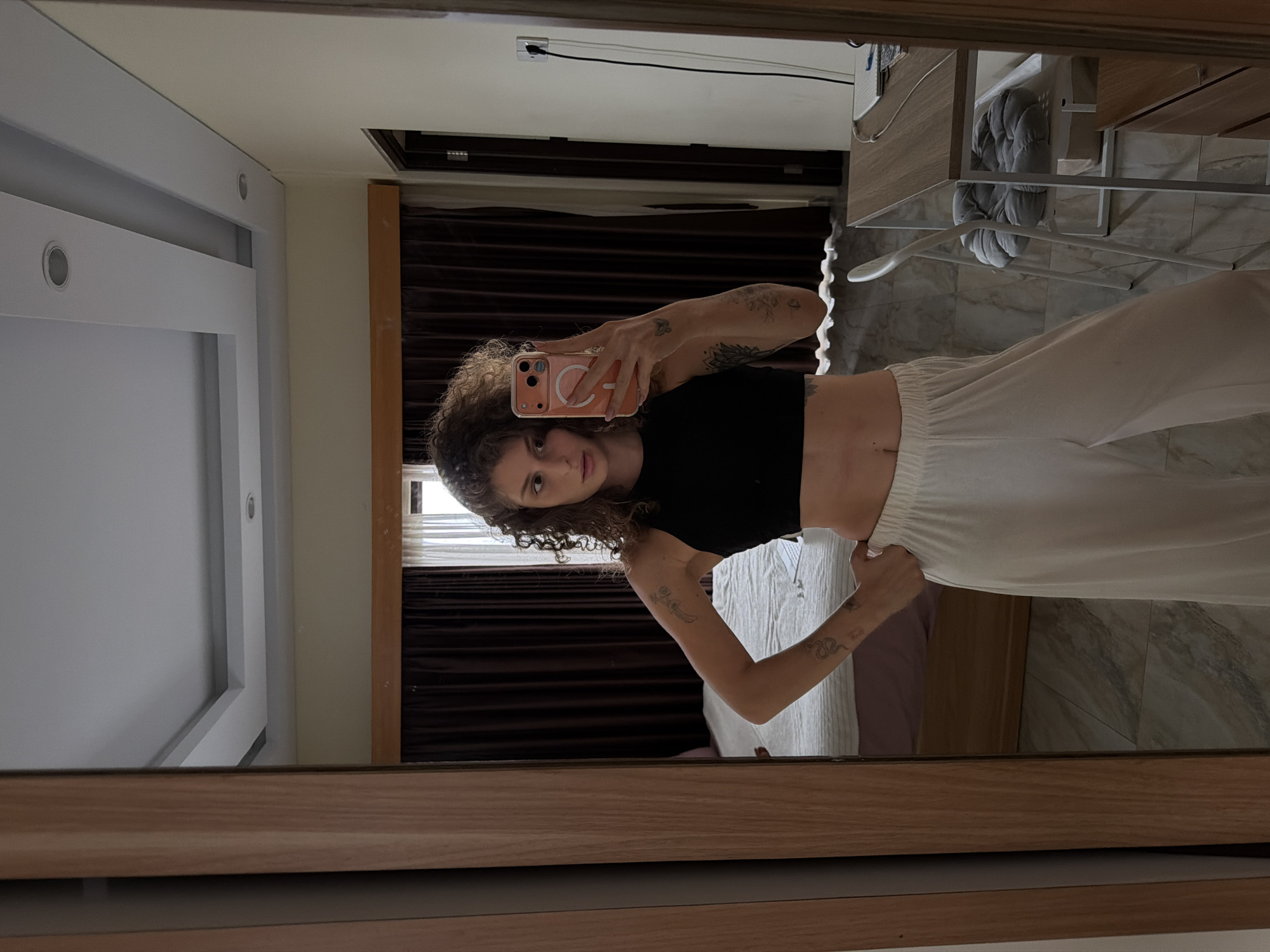 A young woman with curly brown hair takes a mirror selfie in a room. She is wearing a black crop top and white wide-leg pants. She has tattoos on her arms. In the background, a bed with a white comforter is visible, along with dark brown curtains and sheer white curtains. A wooden dresser and a desk with a chair are also in the room. The flooring is a light-colored tile. The room appears to be located in a hotel or guesthouse in North Kuta, Indonesia.