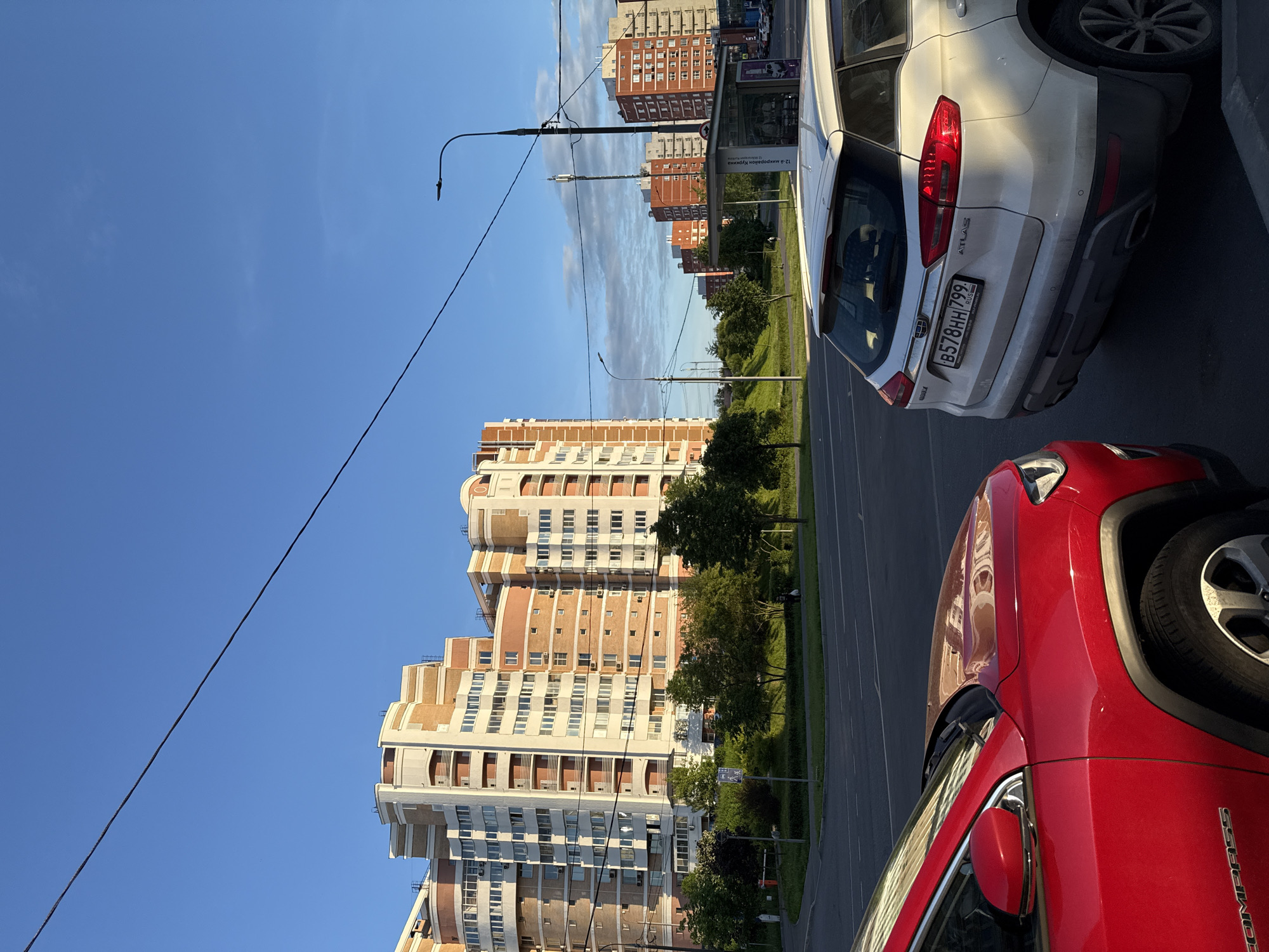 The image depicts an urban street scene on a bright, sunny day with a clear blue sky and a few scattered clouds. The perspective is from street level, looking down a road bordered by green spaces and residential buildings.
In the immediate foreground on the right, the rear of a white Geely Atlas SUV is visible, bearing the Russian license plate "B578HH 799 RUS". The word "ATLAS" is clearly embossed on its tailgate. Partially visible in the bottom right corner is the front-side of a red Jeep Compass SUV, with "COMPASS" visible on its side.
Beyond the parked vehicles, a road stretches into the distance, flanked by a narrow strip of green lawn and mature trees. Several multi-story residential buildings dominate the background. On the left, a large, architecturally distinctive building features a stepped, tiered design with light-colored facades and reddish-brown accents, including numerous balconies. Further down the street and in the distance, other multi-story apartment buildings with more conventional brick-like facades are visible.
Overhead, power lines or tram wires crisscross the sky, supported by tall streetlights. A bus stop shelter is partially visible on the right side of the road, and Russian text on it includes "12-й микрорайон Кунцево" (12th microdistrict Kuntsevo), indicating a location in Moscow, Russia. The bright sunlight creates clear shadows, suggesting either morning or late afternoon. No people or specific activities are depicted, contributing to a tranquil urban atmosphere.
