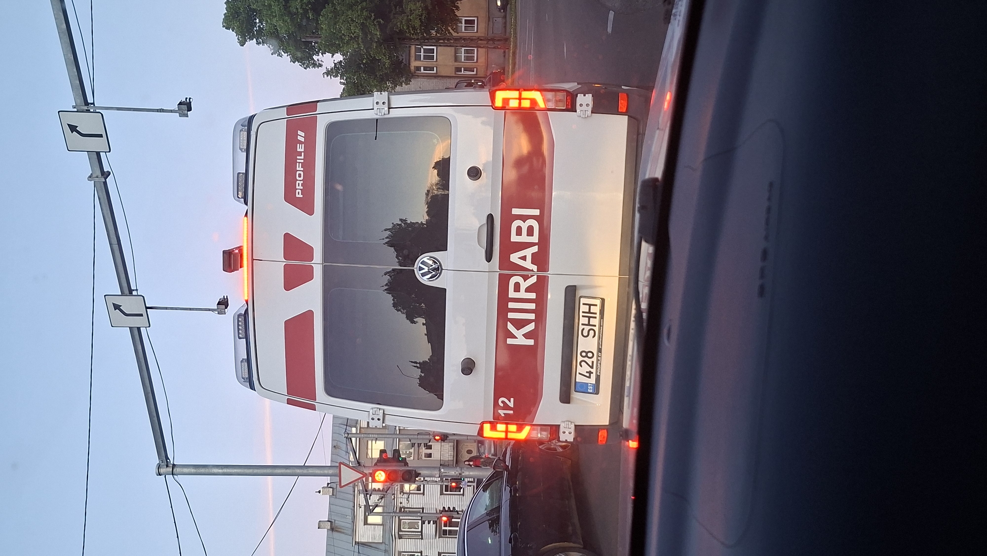 The image shows the rear view of a white ambulance with red stripes and the word "KIIRABI" in red letters. The ambulance has the license plate "428 SHH EST". It is at a traffic light in an urban environment. There are traffic signs overhead indicating a right turn. In the background, there are buildings and trees. The sky is dusky, suggesting it is either dawn or dusk. The scene appears to be a street intersection in Tallinn, Estonia, likely a relatively busy one given the traffic infrastructure.