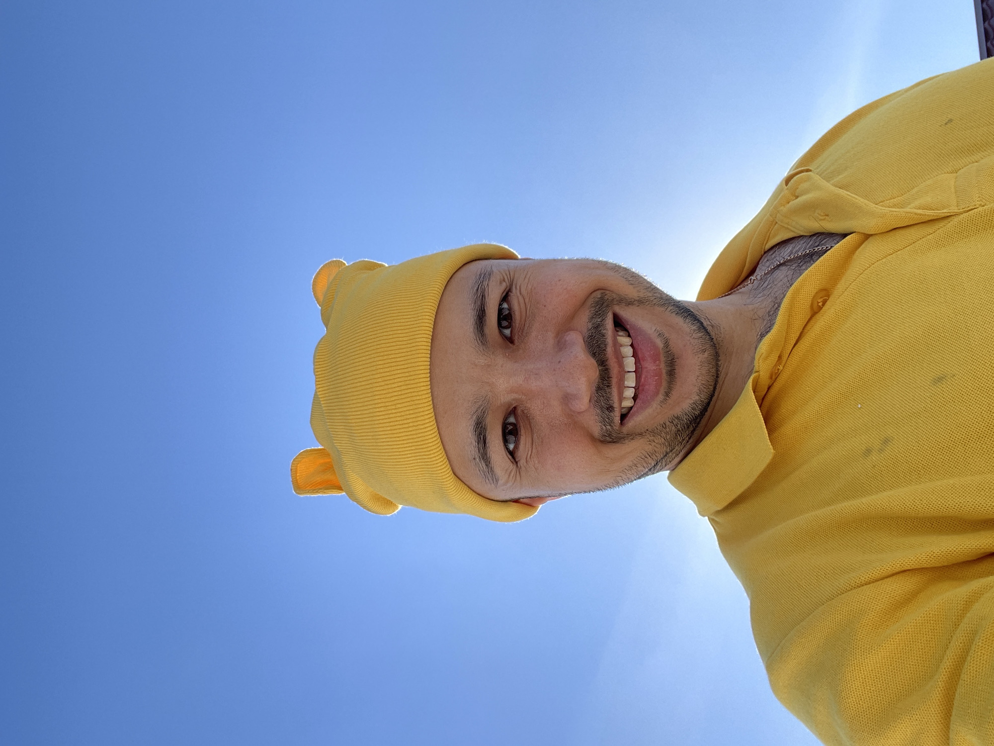 A man with a beard and mustache wearing a yellow bear-eared beanie and a yellow shirt looks down at the camera from an upward angle. The background is a clear blue sky, with the sun shining behind him. The location is unidentifiable as Zav'yalovo, Russia.