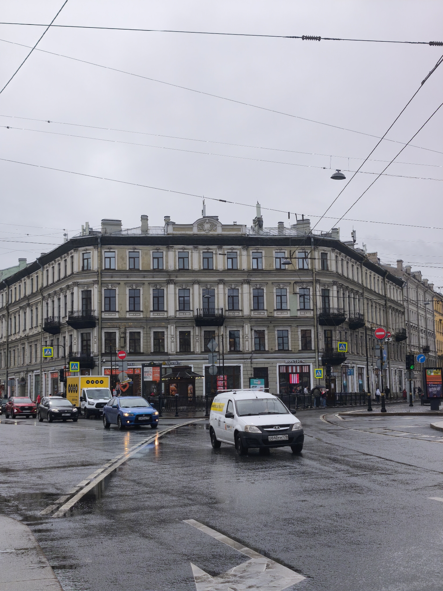 Here's a detailed description of the image:
The image captures a street scene in what appears to be a European city, possibly Russia given the license plates and some of the storefronts. The weather is overcast and wet, with the road surface glistening with rain.
**Setting:** The location is at a busy intersection. The architecture is distinctly European, with a large, ornate building dominating the background. This building has multiple stories, rows of windows, and decorative elements like balconies. There are businesses on the ground floor of the building, including what appears to be a "Rostic's" fast-food restaurant.
**Traffic and Vehicles:** Several vehicles are visible on the road. There's a blue car, a white van with yellow markings, a black car, and a red car. The vehicles are driving on the right side of the road. The white van is prominently featured in the foreground.
**Pedestrians and Street Elements:** There are a few pedestrians visible on the sidewalks, mostly near the storefronts. Traffic lights and pedestrian crossing signs are present. There's also street furniture like lampposts and railings.
**Weather and Environment:** The overall atmosphere is gray and damp. Reflections from the wet road surface add to the sense of a rainy day. Overhead, there are numerous power lines crisscrossing the sky.
**Additional Details:**
* There are arrow markings on the road indicating traffic direction.
* Storefront signs in the windows include the word "fresh" and a price of "49 P" likely referring to Russian currency, Rubles.
* License plates on the vehicles are Russian in style.
