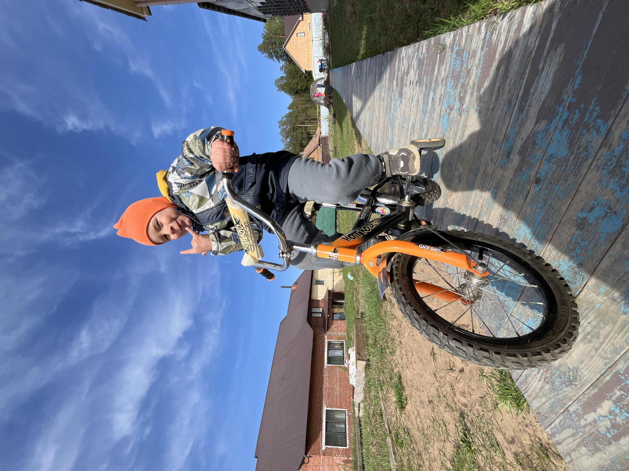 A child on a bicycle is oriented with their head pointing to the upper left of the frame. The child is wearing an orange hat and a jacket with blue and white geometric patterns and a yellow collar. They extend their left hand, making a "hang loose" gesture. The bicycle is black and orange. The background includes a bright blue sky with wispy clouds. A wooden walkway runs from the middle to the upper right, with patches of blue paint. Beyond the walkway is grass and foliage, and two buildings are visible in the background. One is a brick structure with a dark roof, and the other is a light brown building. Based on the location information, this scene likely takes place outdoors in the city of Chaikovsky, Russia.