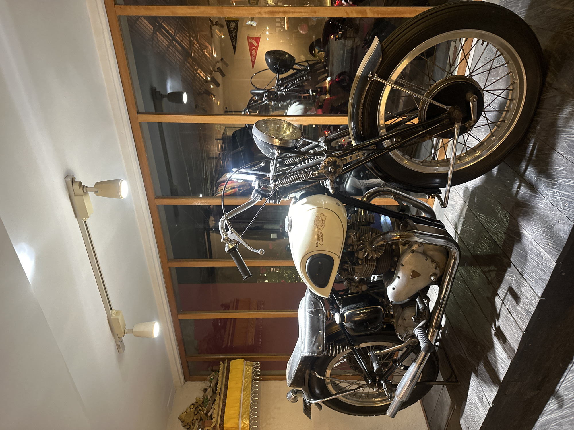 A vintage motorcycle is on display indoors in Ubud District, Indonesia. The motorcycle is a light color with dark fenders and spoked wheels. The location appears to be a gallery or shop, with a mirrored wall reflecting other motorcycles and interior details. The lighting is bright, and the environment seems well-maintained.