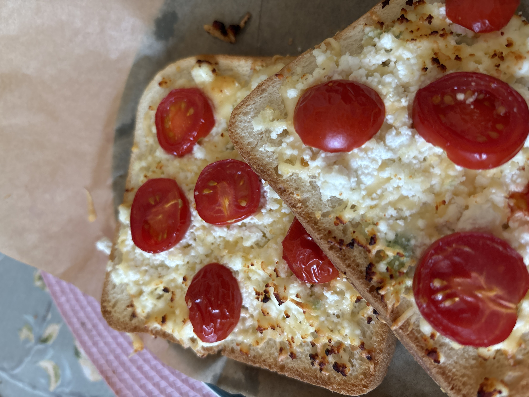 The image shows two slices of toast topped with crumbled cheese and cherry tomatoes. The toast is golden brown and appears to be slightly toasted. The cheese is white and crumbly, and some of it has melted and browned in places. The cherry tomatoes are red and appear to be halved or quartered. They are scattered across the cheese. The toasts are arranged on a piece of parchment paper, which is on a patterned surface. The background is blurred but appears to be a table with a pink placemat. The image is taken from a close-up, overhead perspective. There is no information available about the location in Naberezhnye Chelny, Russia.