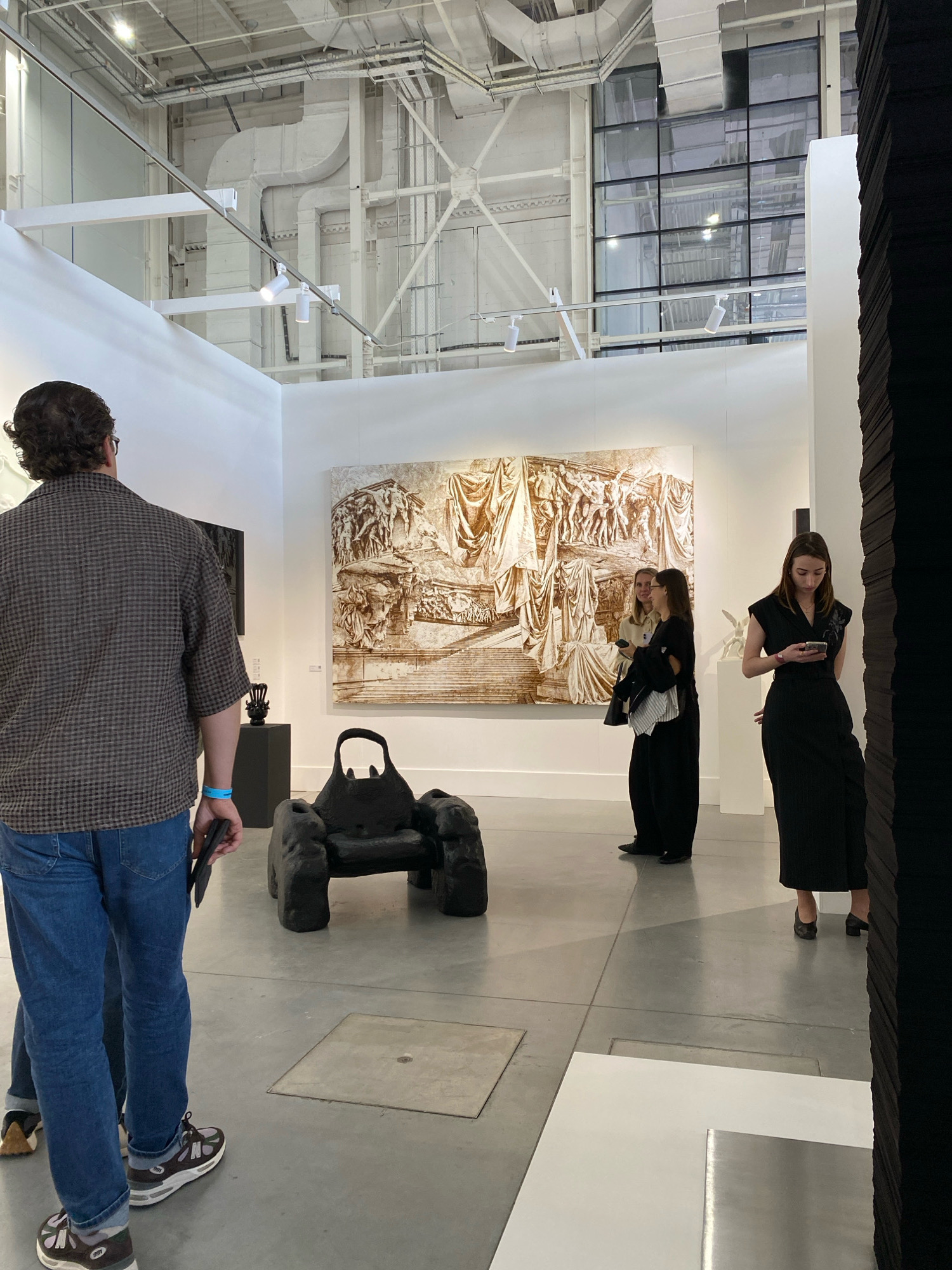 The image depicts an indoor art gallery. In the foreground, a man with dark curly hair and glasses, wearing a plaid shirt and blue jeans, is viewed from behind, holding a tablet. To his right is a dark, amorphous sculpture resembling a chair. Further into the gallery, a large, monochromatic artwork with classical figures is displayed on a white wall. Three individuals are interacting near this artwork: two women in dark clothing are facing the artwork and conversing, while another woman in a black dress is looking at her phone. The gallery space features polished concrete floors, white walls, and exposed industrial ceiling elements with lighting fixtures. To the right, a tall, dark, textured art installation is partially visible. The overall environment suggests a contemporary art exhibition. The location is identified as Khimki, Russia.