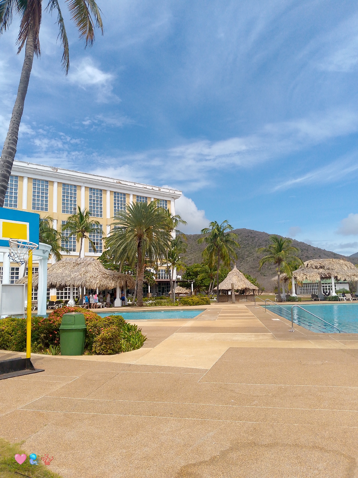 This image captures a sunny day at a resort in Pedro González, Venezuela. A large, yellow and white hotel building with multiple stories is visible in the background, partially obscured by palm trees. In front of the hotel are swimming pools separated by a walkway made of light-colored concrete. Thatched-roof structures are scattered around the pool area, likely for shade and relaxation. Some people are gathered near the first structure, while others appear to be relaxing or swimming in the pools. A basketball hoop is positioned near the edge of one pool, beside a bush-lined area and a green trash can. The landscape includes a mountain in the distance, under a blue sky with scattered clouds.