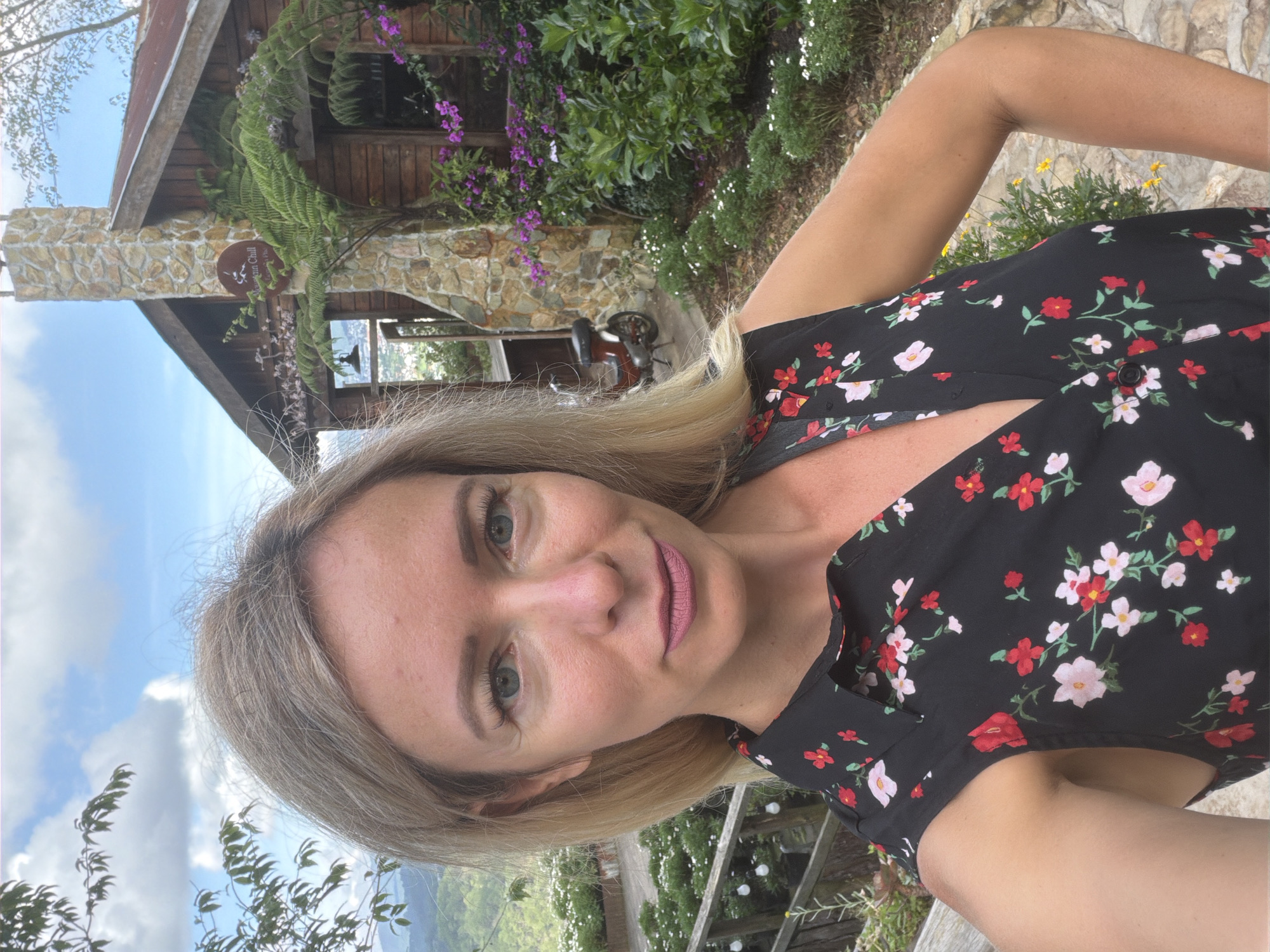 An outdoor selfie features a Caucasian woman with blonde hair and blue eyes, wearing a black floral dress. She is positioned in the foreground, looking directly at the camera. Her left arm is bent at the elbow, and her hand is resting on her hip. Behind her, a wooden cabin with a stone chimney and a red-tiled roof is visible, surrounded by lush greenery and flowers. A small red scooter is parked next to the cabin. The scene is set against a backdrop of rolling hills and a cloudy blue sky. The overall setting appears to be a rural or mountainous area, potentially a resort or a private residence in Xuân Thành, Vietnam.