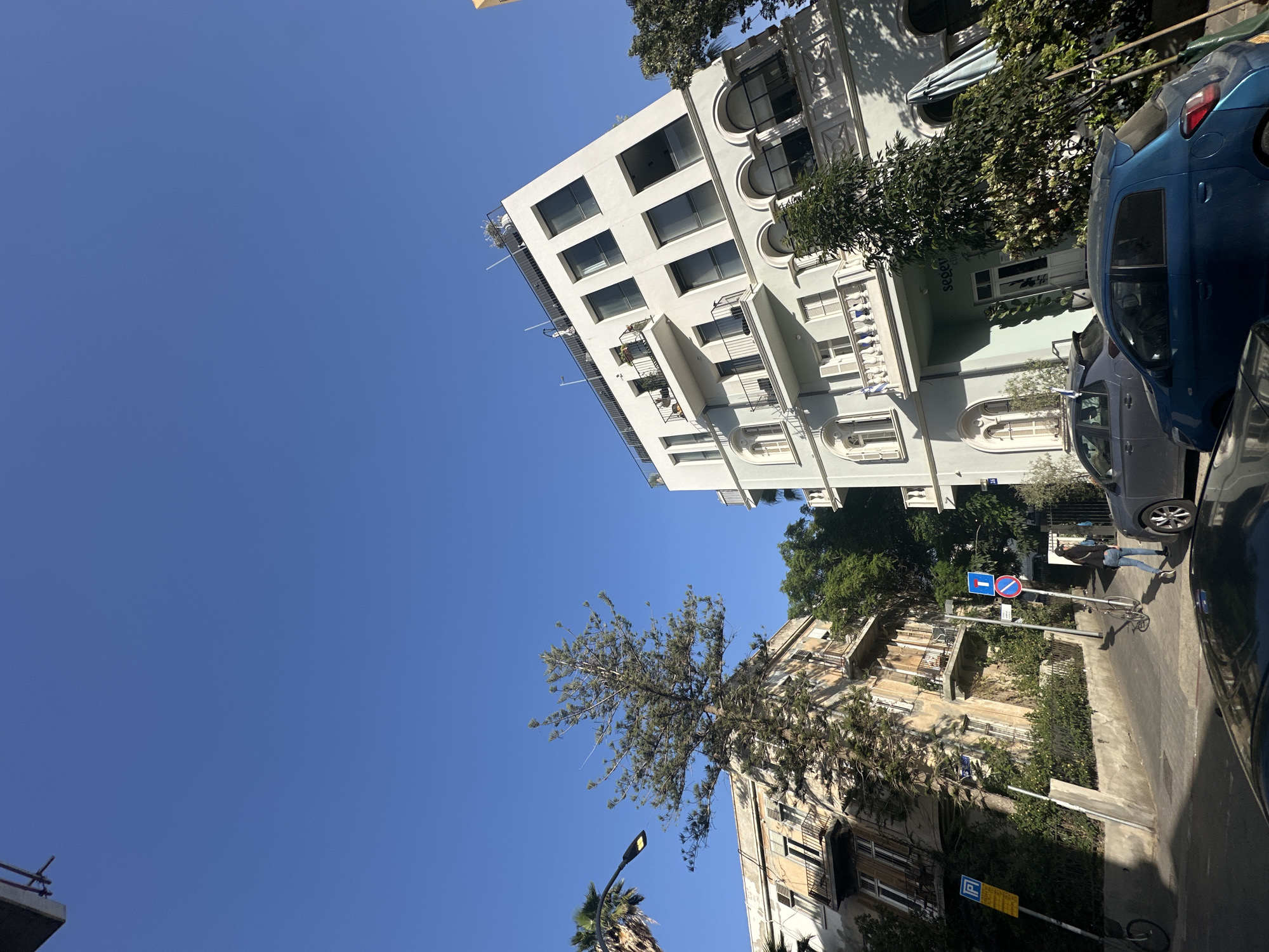 The image shows a street scene in Tel Aviv, Israel, taken on a sunny day. The camera is pointed upward, capturing a view of buildings against a clear blue sky.
On the right side of the frame, a multi-story white building with a modern design is visible. It features a symmetrical facade with rows of windows and balconies. Adjacent to the white building is a section of a parked blue car.
To the lower right, another building is seen. This building has a more traditional style. A tree stands in front of this building.
Street signs, and other infrastructure are also visible. A person is walking on the sidewalk.