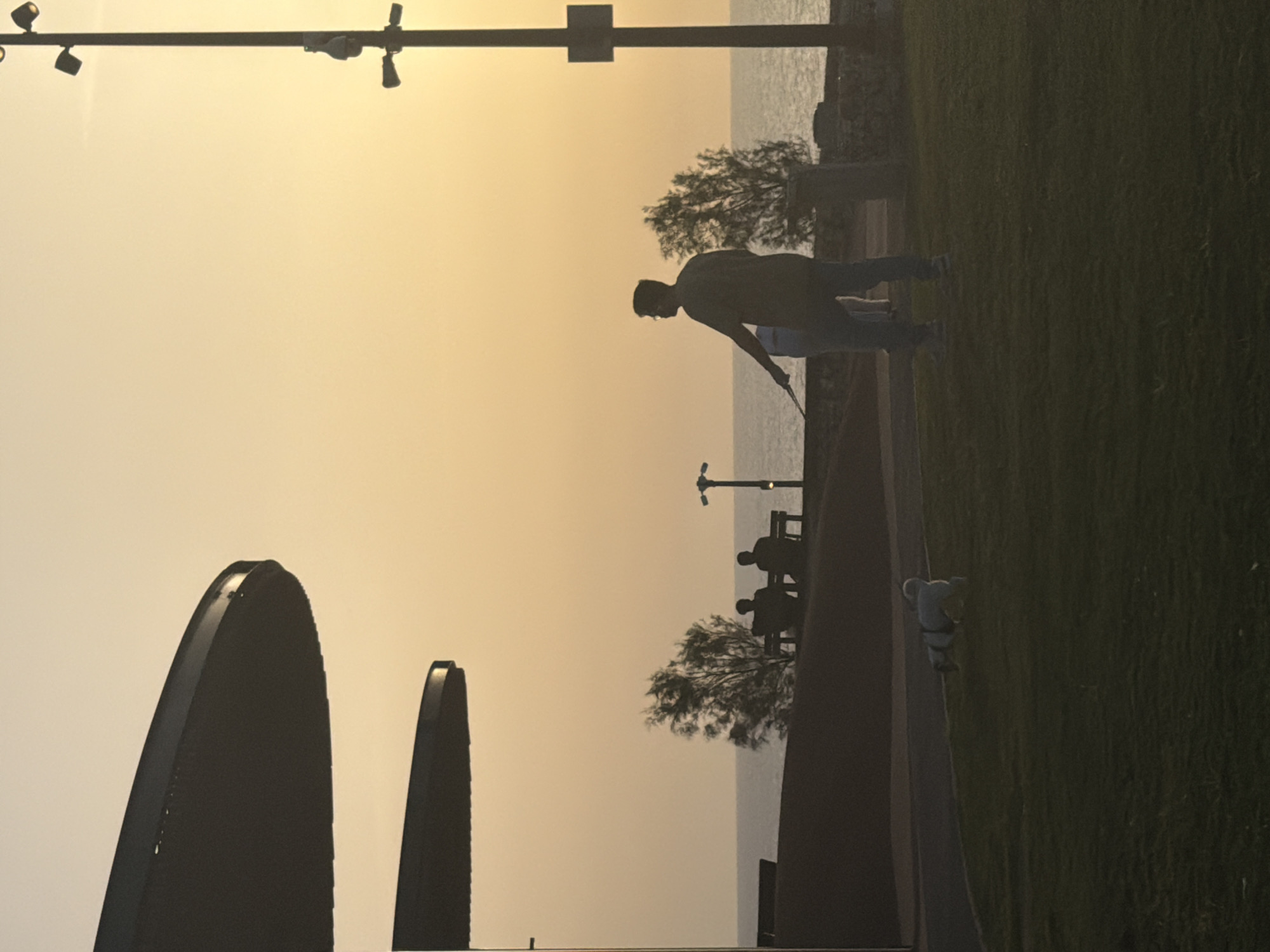 The image depicts a coastal scene in Rishon LeZion, Israel. It captures a man walking on a path along the coast with a dog. In the background, there are two individuals sitting on a bench. The lighting suggests it is sunset or sunrise. The scene includes modern structures in the foreground.