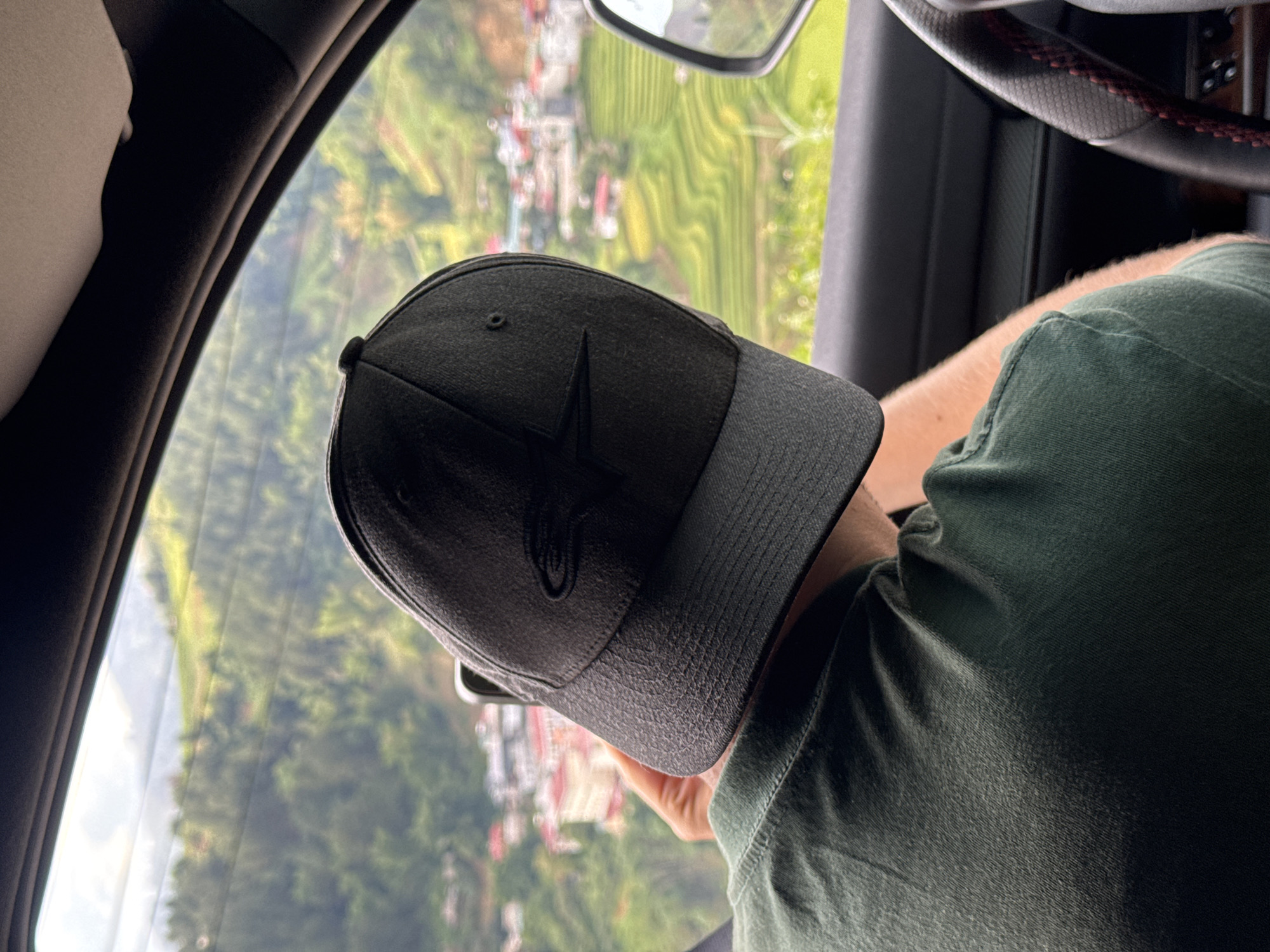 The image depicts a person viewed from behind and slightly to the side, seated inside a vehicle and looking out a side window at a landscape in Khao Mang, Vietnam. The person wears a dark green short-sleeved t-shirt and a dark grey/black baseball cap. A black, stylized star logo, characteristic of the Alpinestars brand, is visible on the front of the cap.
The interior of the vehicle is partially visible, including a beige headrest or seat cushion on the left, and a steering wheel with distinctive red stitching on the right, suggesting it's a car. The individual's arm rests near the steering wheel as they gaze outside.
Through the car window, a vibrant, sunlit landscape is visible, though somewhat blurred, indicating motion or a shallow depth of field. The scene features rolling green hills or terraced fields, possibly for agriculture like rice or tea, interspersed with small buildings or houses. This suggests a rural or mountainous region, consistent with the provided location context of Khao Mang, Vietnam. The time of day appears to be daytime with clear weather. No other discernible text is present.
