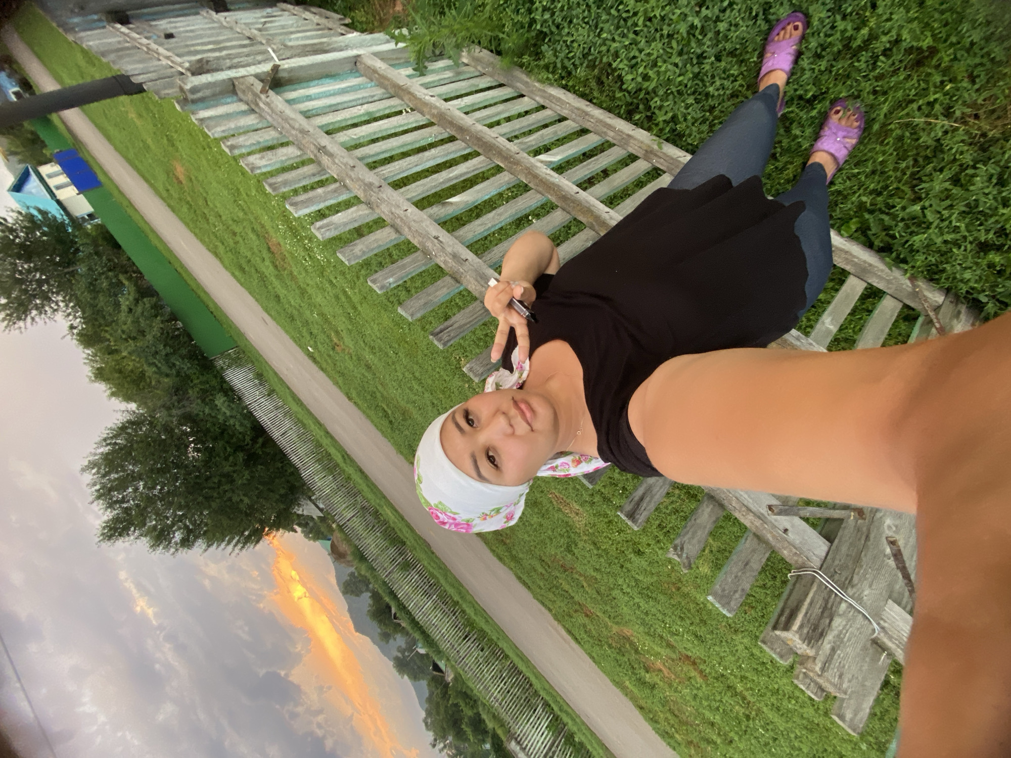 In Maloarslangulovo, Russia, a woman with a white and pink floral headscarf and a black top poses for a selfie by a weathered wooden fence. She is wearing blue jeans and purple fuzzy slippers. Behind her, a paved road runs diagonally through a grassy area, bordered by fences and trees. In the distance, houses and a mountainous landscape are visible under a dramatic sunset sky with orange and gray clouds.