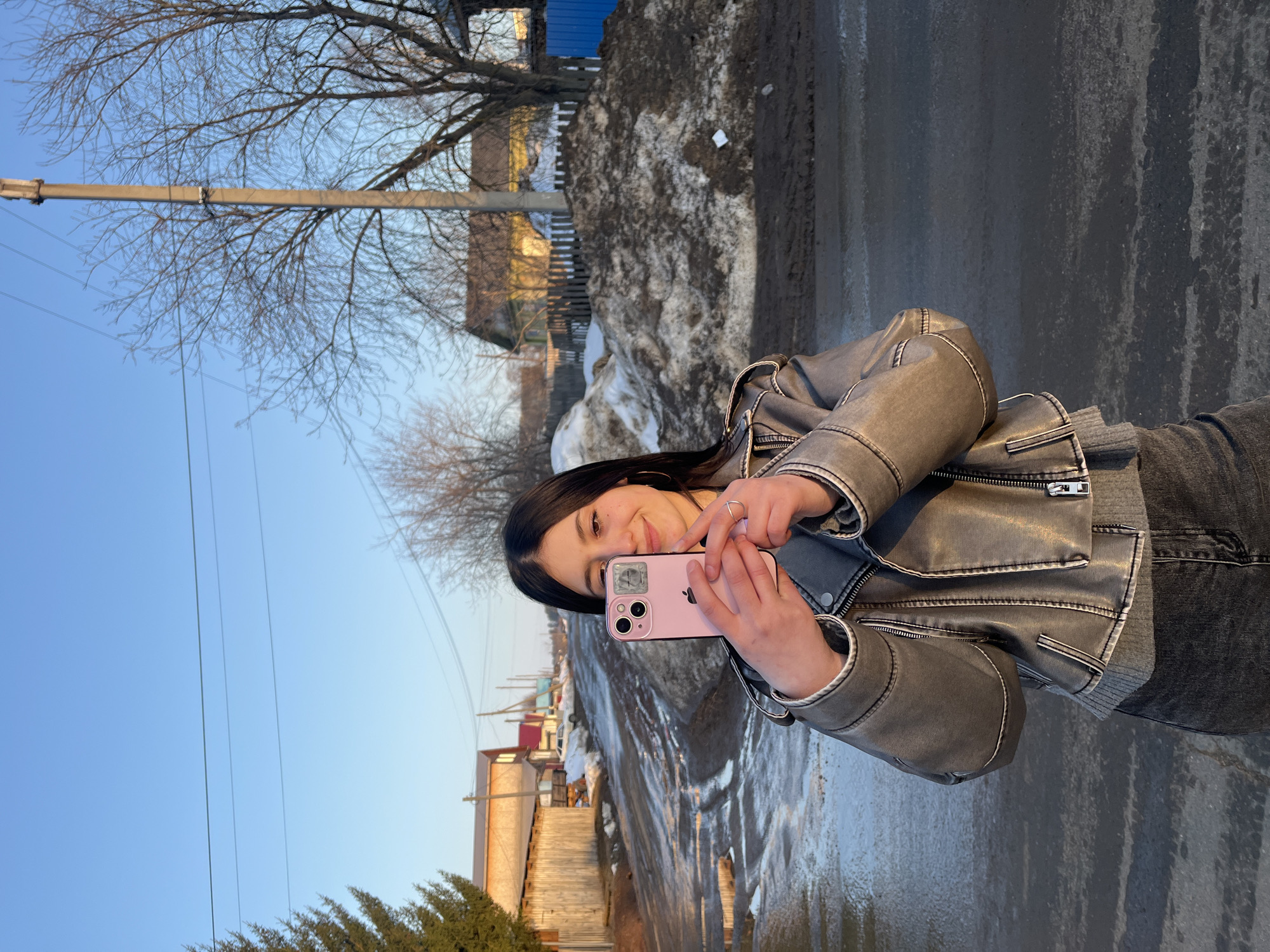 A young female, appearing to be in her late teens or early twenties, stands centered slightly to the right of the frame on a muddy dirt road in the city of Фоки, Russia. She has long dark hair and is smiling, looking towards a pink iPhone that she holds horizontally with both hands, positioned in front of her face as if taking a selfie or recording video. The phone displays a reflective sticker or object, possibly a condom wrapper, over the top left camera lens. She wears a distressed grey faux leather jacket over a grey sweater and dark jeans. A silver ring is visible on her right hand.
The environment depicts a rural or semi-rural street during a period of thaw, likely early spring. The unpaved road is uneven, wet, and muddy, with visible puddles and patches of melting ice or snow. To the left, a wooden fence runs alongside the road, followed by an evergreen tree. Further down the street, various single-story houses, some with light-colored or red roofs, are visible. To the right, large, irregular piles of dirty snow and mud line the roadside. Behind these piles, a wooden fence, a house with a yellow exterior, and a section of blue corrugated metal fencing are visible. Bare deciduous trees are scattered throughout the background. Overhead, a clear blue sky is punctuated by power lines extending from a concrete utility pole. The lighting suggests late afternoon or early morning sun.