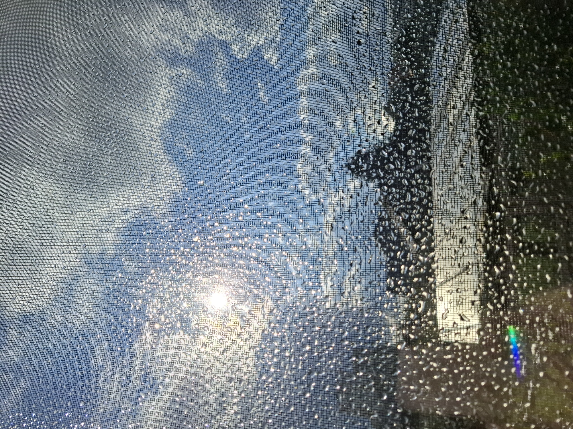 The image depicts a view through a window screen covered in water droplets. The background shows a bright, sunny sky with scattered clouds. Sunlight shines through the water droplets, creating a sparkling effect. In the distance, a dark, silhouetted landscape with a body of water is visible. The overall impression is a somewhat abstract and textured view of an outdoor scene, likely a cityscape, seen through a wet window. There is no specific visual information within the image that indicates it is located in Tyumen, Russia.