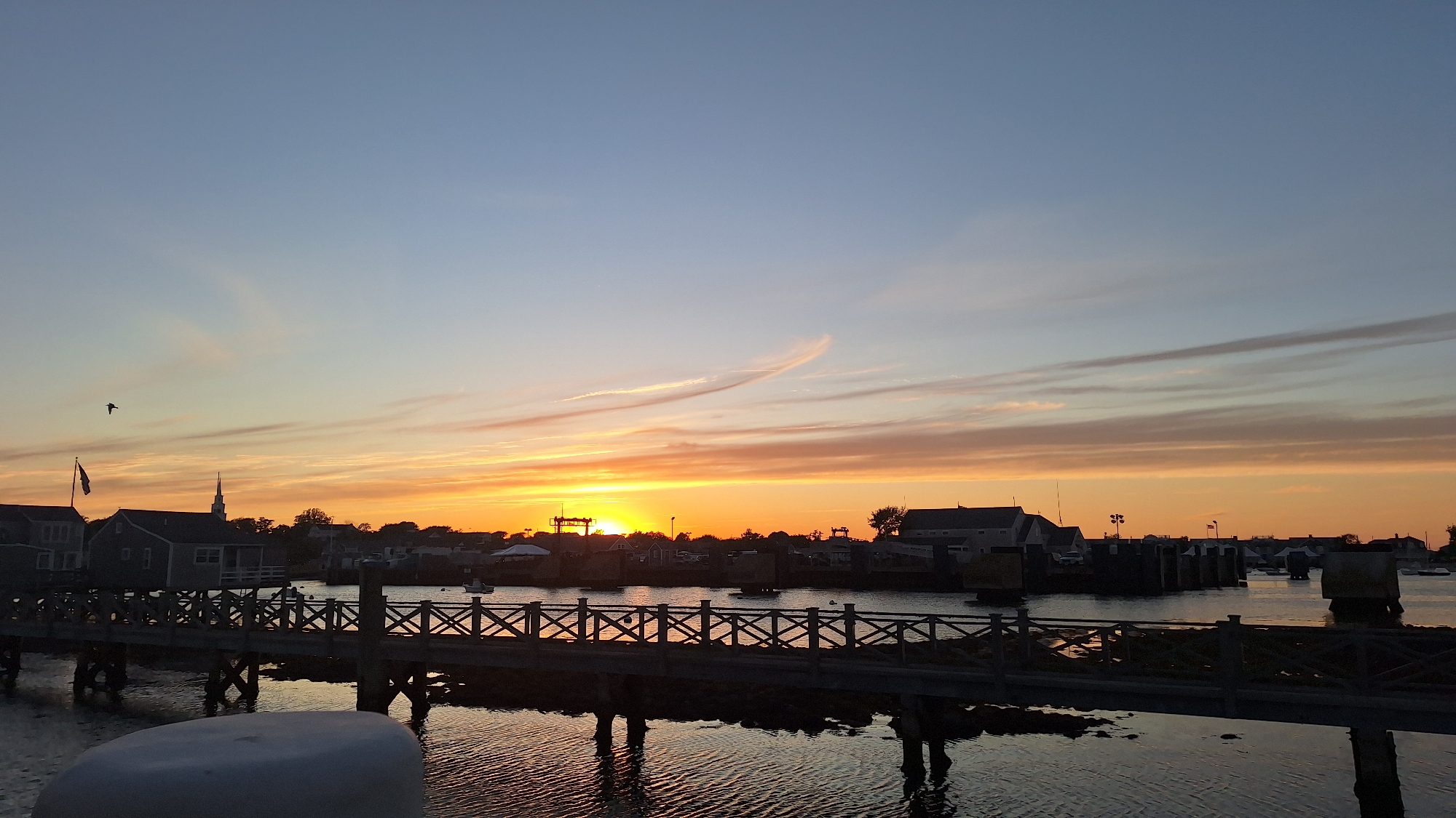 A silhouette of a wooden boardwalk with cross-hatching railing is in the foreground, spanning over dark, reflective water. Beyond the boardwalk, a sunset casts an orange and yellow glow across the horizon, illuminating thin, wispy clouds in a pale blue sky. Several buildings, including a white steeple, are visible along the shoreline, silhouetted against the sunset. Boats are docked in the water. A bird is in flight in the upper left quadrant of the image. The scene appears to be a harbor at sunset, likely on the city of Nantucket in the United States of America, given the architecture and waterfront setting.