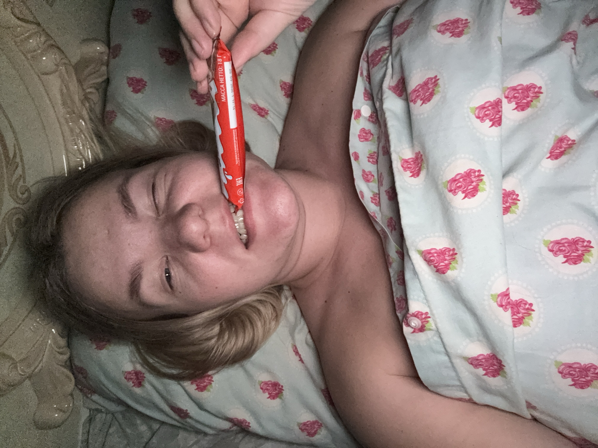 A woman lies in bed, seen from a high angle, appearing to take a selfie or be photographed by someone beside her. She has fair skin and blonde hair, and is wearing a light blue garment with a pink rose pattern, matching the duvet or comforter she is partially covered by. Her left eye is mostly closed, and her nose is crinkled as she smiles, showing her teeth, while holding a red candy bar to her mouth. Her right arm is visible, bare.
The setting appears to be a bedroom, with a decorative light-colored headboard visible on the left side of the frame. The lighting is dim, suggesting it might be evening or night. On the red candy wrapper, the text "МАССА НЕТТО: 18 г" (NET WEIGHT: 18 g) is clearly visible, along with the "Kinder" brand logo. The woman's expression conveys enjoyment or playful satisfaction. The scene is set in Chaikovsky, Russia.