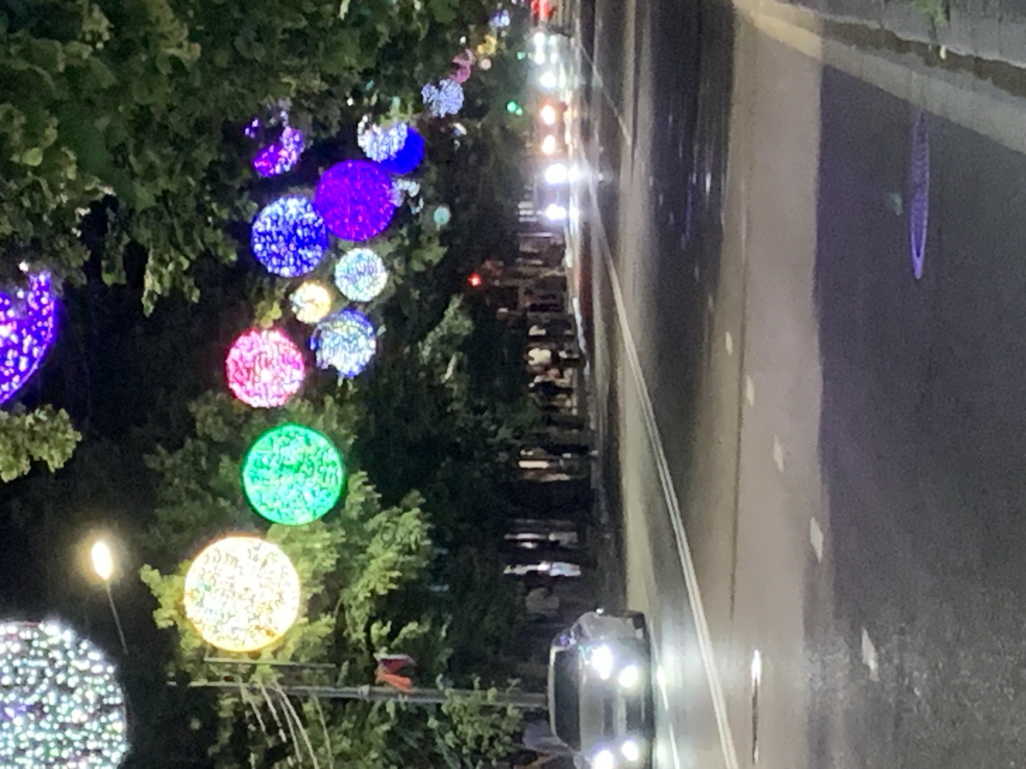 The image shows a nighttime street scene in an unspecified city. Large, illuminated spherical decorations hang from trees lining the street, casting colorful lights. A silver car with its headlights on is driving down the road, and other vehicles are visible in the distance. Pedestrians are walking on the sidewalk, partially obscured by trees. The road is paved with asphalt and has white lane markings. Some areas of the road appear wet, reflecting the lights. A street lamp is visible in the upper left portion of the image.