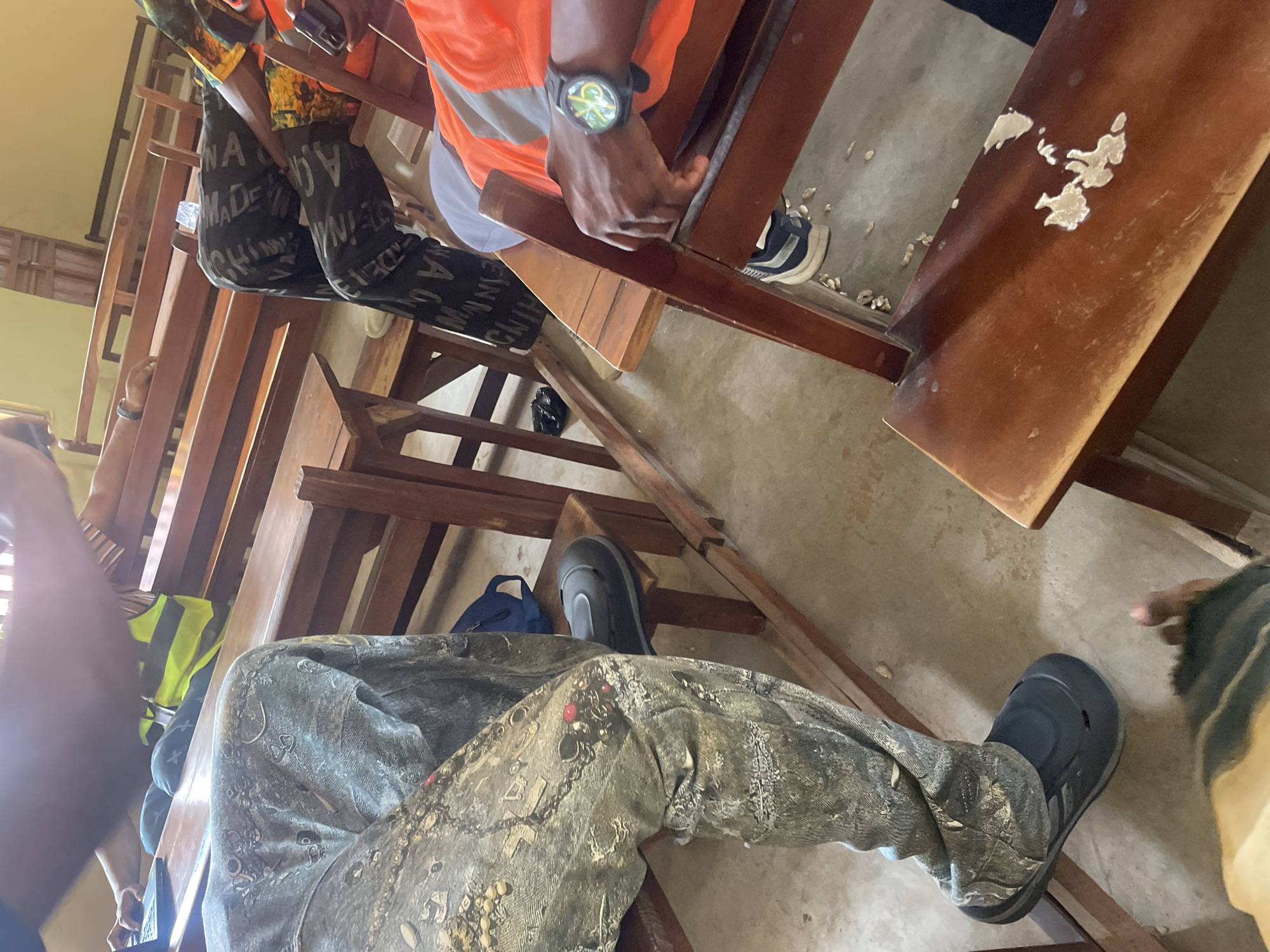 The image shows several people sitting on wooden benches in what appears to be a classroom or hall in Lokoja, Nigeria. One person in the foreground is wearing camouflage-patterned pants and dark slip-on shoes. To their right, another individual is wearing black pants with white lettering and an orange and gray safety vest over a colorful shirt. This person is also wearing dark sneakers. A third person is partially visible in the upper left, wearing a bright yellow safety vest and black pants, and is using a laptop. The benches are made of dark wood and are arranged in rows. The floor is concrete and appears to have some debris on it. The lighting suggests an indoor setting, possibly with natural light coming from a window or open area. The overall scene depicts a gathering of individuals, likely for a meeting, training session, or educational activity.