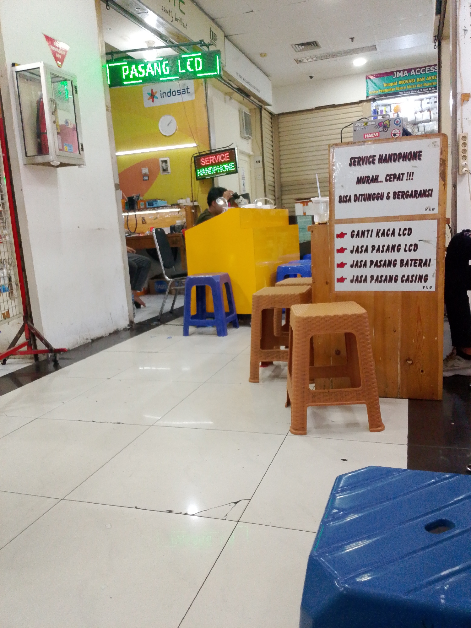This image shows a section of a commercial area in Central Jakarta, Indonesia. In the foreground, a bright blue plastic stool is prominently featured. The floor is tiled with white square tiles, some of which show signs of wear and cracking.
To the left, a white wall meets a tiled floor and a metal display rack. Above the wall, a green LED sign reads "PASANG LCD" which translates to "INSTALL LCD". Below this sign, the Indosat logo is visible.
In the mid-ground, a yellow counter-like structure is present, behind which a person is partially visible. A blue plastic stool is to the left of the yellow structure. Further right, a wooden stall is set up with a sign that says "SERVICE HANDPHONE" (HANDPHONE SERVICE). This sign lists services such as "GANTI KACA LCD" (REPLACE LCD GLASS), "JASA PASANG LCD" (LCD INSTALLATION SERVICE), "JASA PASANG BATERAI" (BATTERY INSTALLATION SERVICE), and "JASA PASANG CASING" (CASING INSTALLATION SERVICE). Several brown plastic stools are placed in front of this wooden stall.
In the background, a banner for "JMA ACCESS" is displayed. The overall environment suggests a retail or service center focused on mobile phone repairs and accessories.