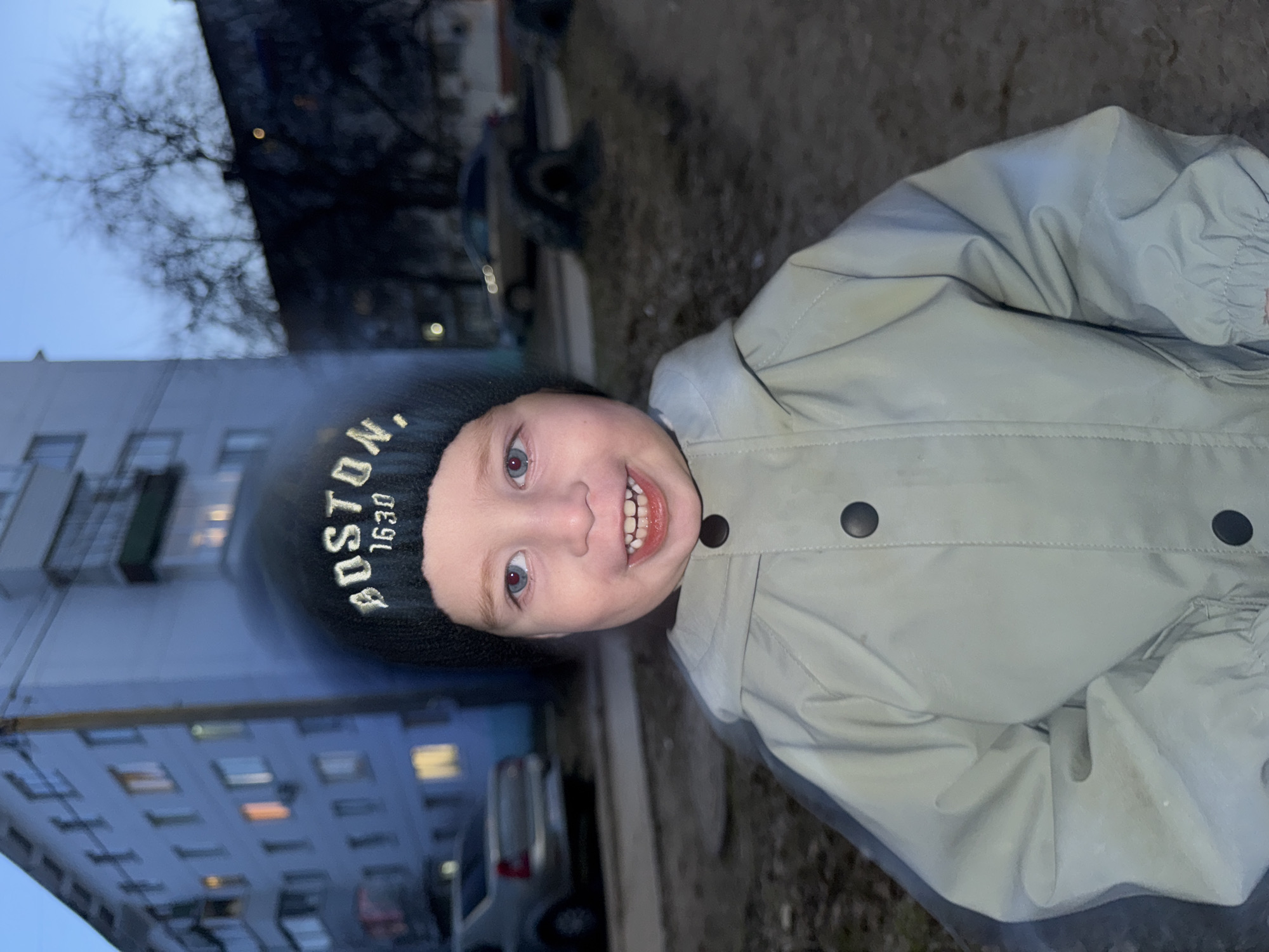 A child with fair skin, red eyes, and a wide grin is wearing a black beanie with "BOSTON 1630" embroidered on it, and a gray button-up jacket. The background includes a multi-story building with lighted windows, bare trees, vehicles, and a sidewalk. The image appears to have been taken at dusk or in low light. Based on the building architecture and vehicles, the location is likely a residential area in the city of Chaikovsky, Russia.
